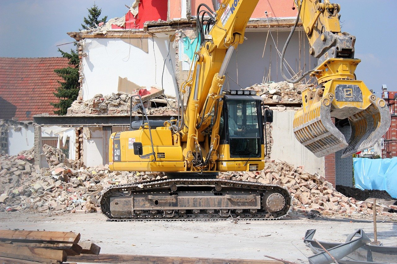 A yellow excavator tearing down a building. Debris and rubble are scattered around in the demolition site.