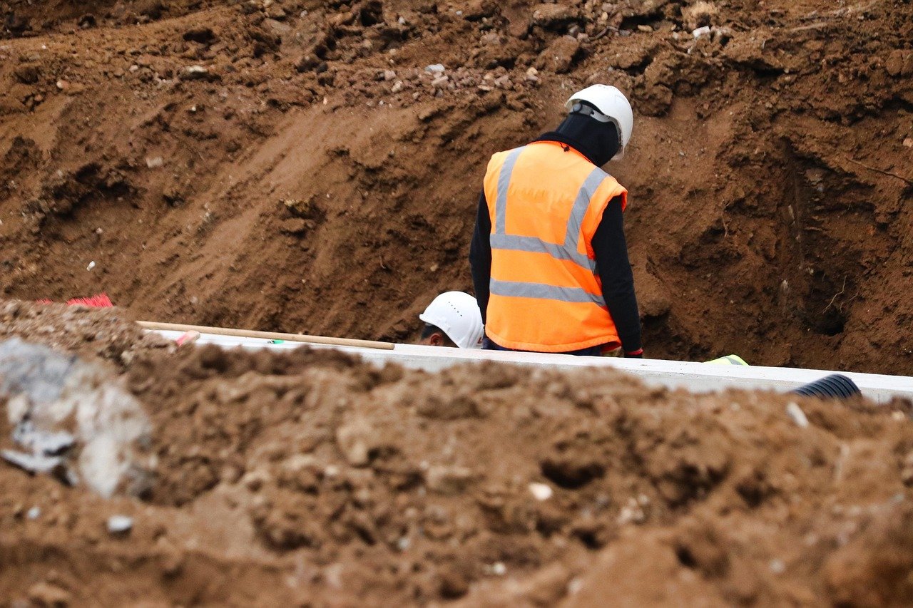 Construction workers in safety gear working on a construction site with an open trench and dirt surrounding them.