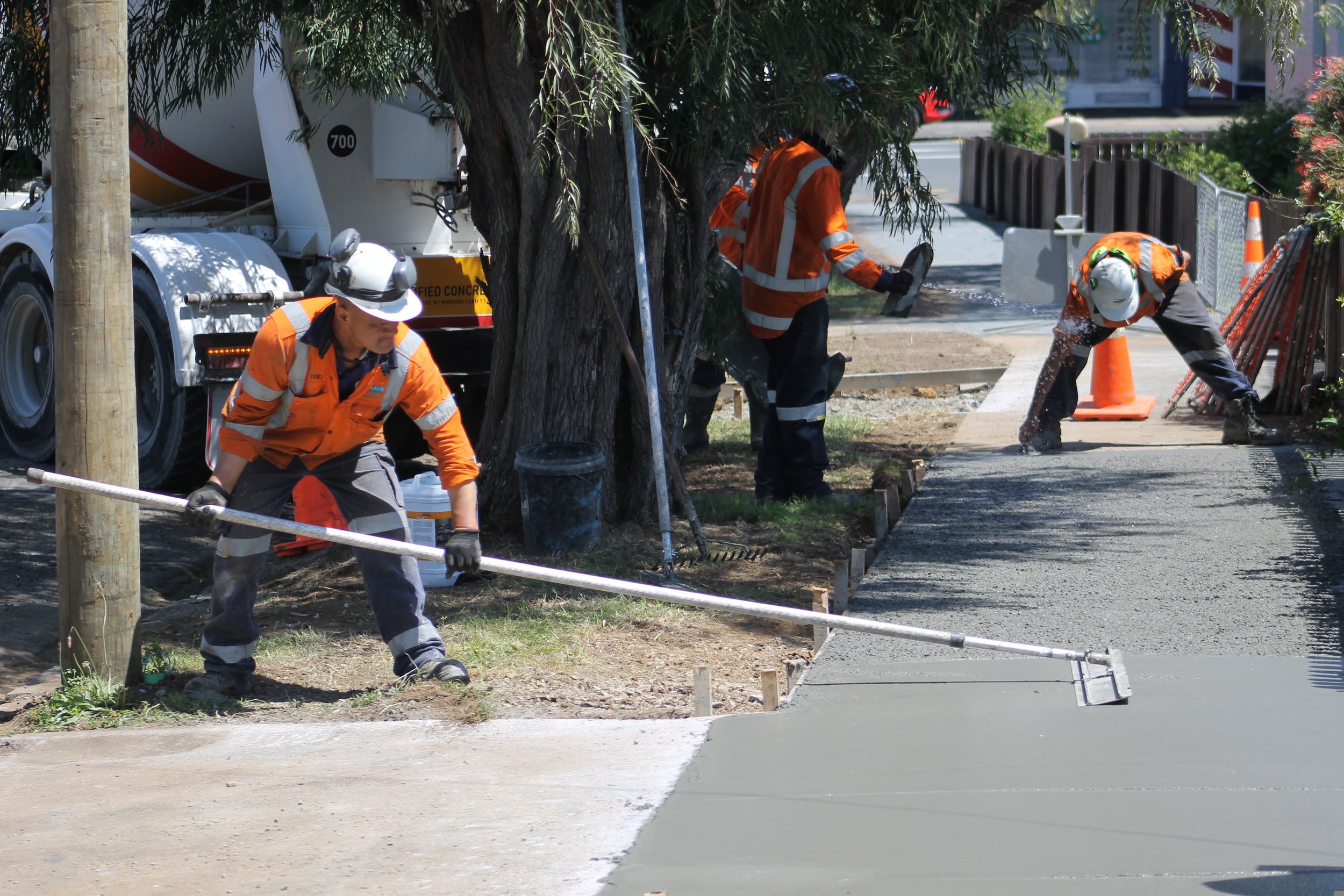 Construction workers paving a sidewalk with concrete, wearing orange safety vests and helmets, using tools and equipment.
