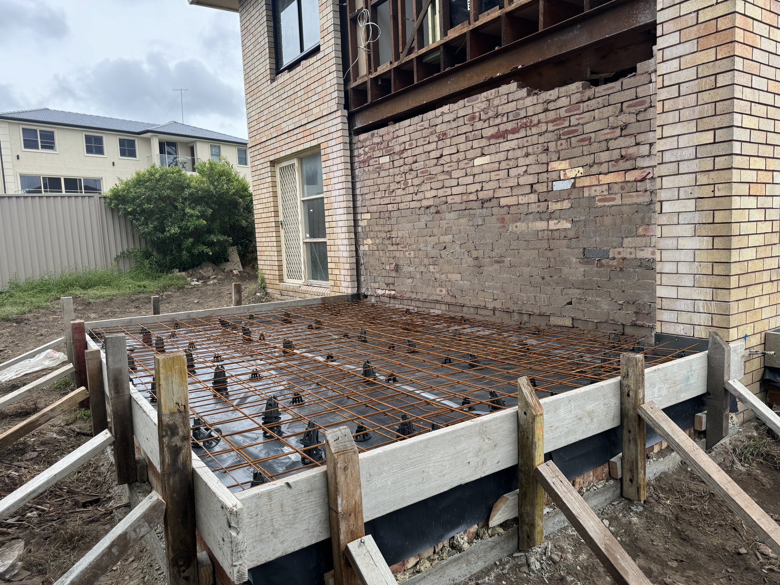 Construction site showing a concrete slab with rusted rebar and formwork around the edges next to a brick house with a sliding door and garden area.