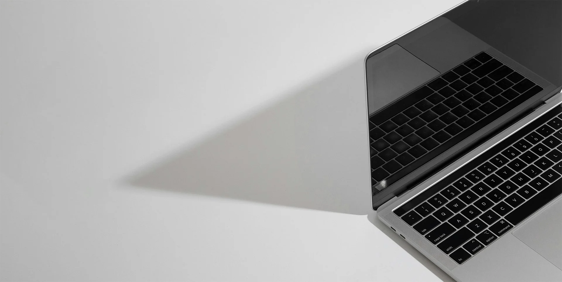 Top view of an open silver laptop with a black keyboard on a white surface, casting a shadow.