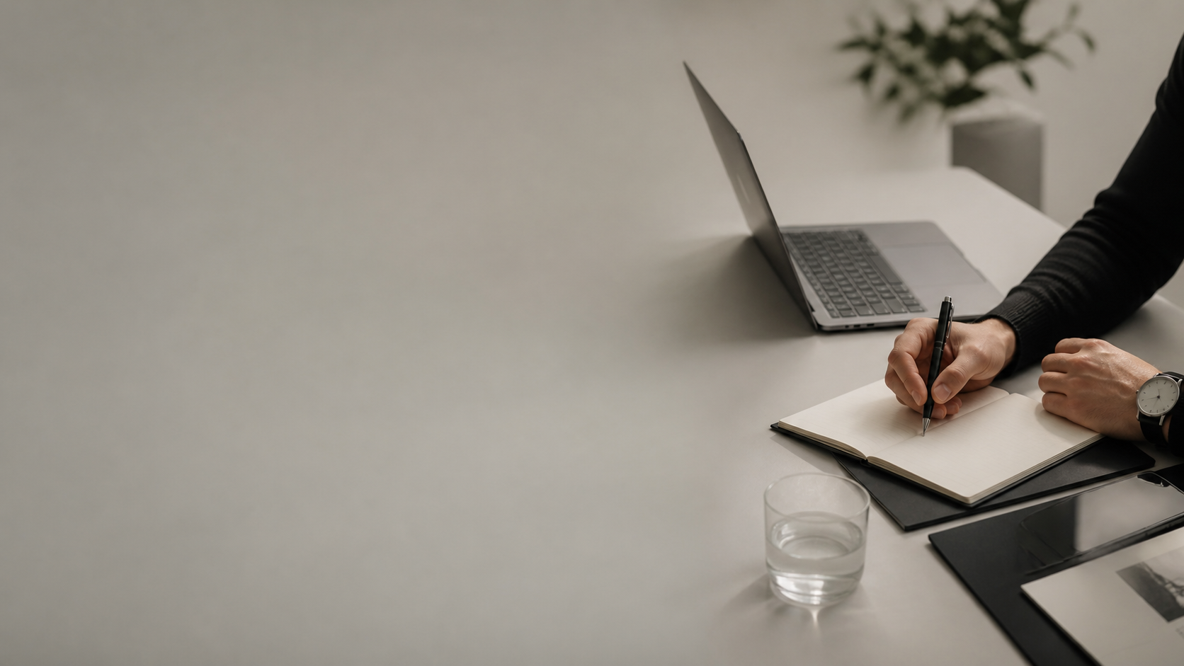 Person writing in a notebook at a white desk, with a laptop, glass of water, and some documents nearby.