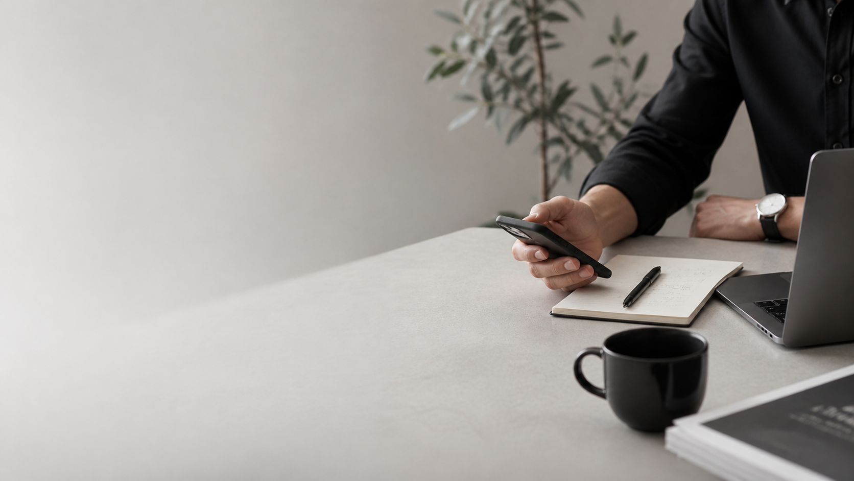 Person in black shirt sitting at desk with a laptop, smartphone, notebook, pen, and black coffee mug.