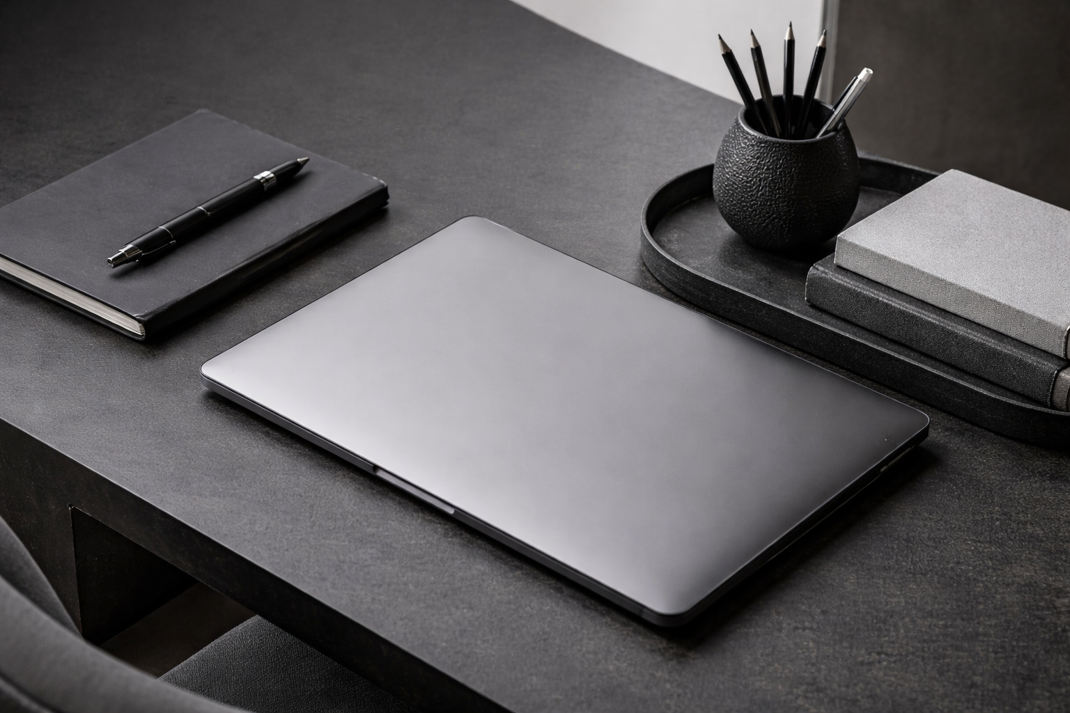 A closed silver laptop, black notebook with a pen, and a black textured vase with pens, on a dark desk with gray and black books and a black tray.