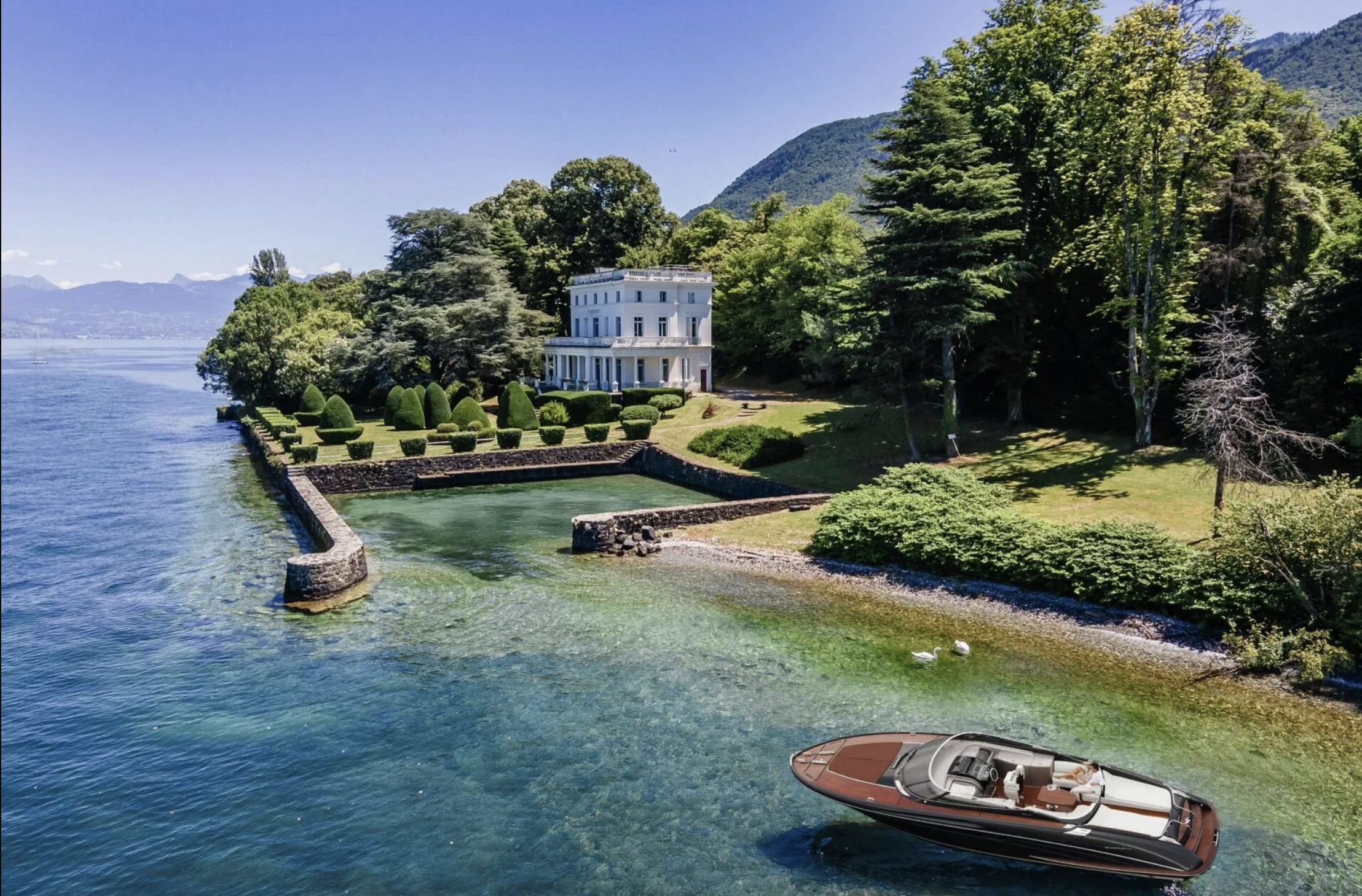 A lakeside mansion with manicured lawns, a stone wall, and a boat on the water, surrounded by lush trees and mountains in the background.