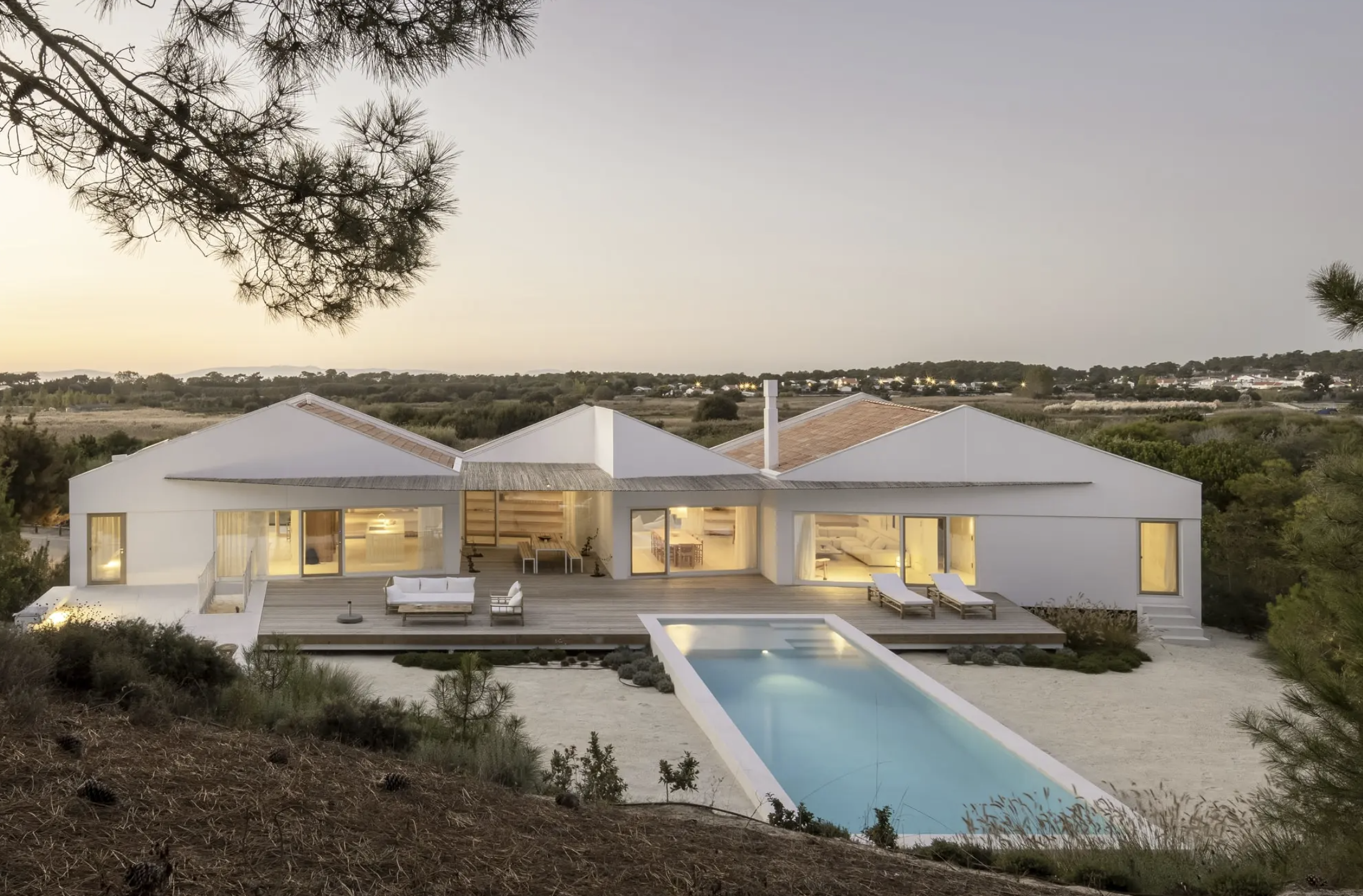Modern white house with large windows, a wooden deck, outdoor seating, a rectangular swimming pool, and surrounding natural landscape at dusk.
