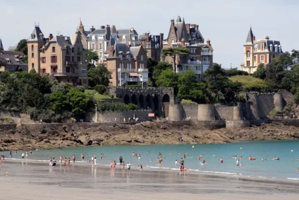 Beach scene with people swimming and playing in the water, rocky shoreline, and stylish houses on a green hill in the background.