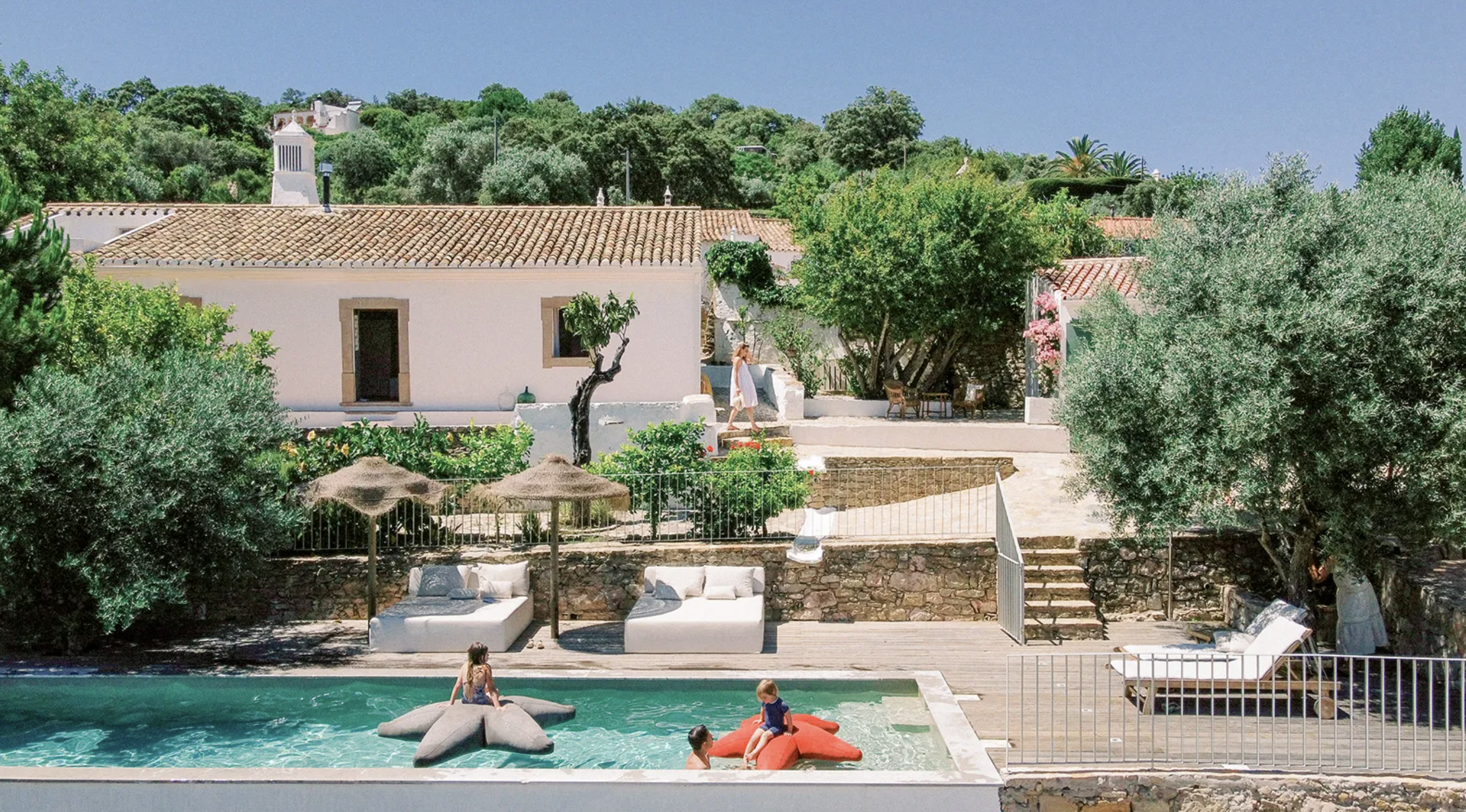 Children playing on inflatable star and flower floats in a swimming pool at a private outdoor patio. The patio has lounge chairs, umbrellas, and lush greenery including trees and bushes. A woman in a white dress walks near the pool area, with a backdrop of white houses with tiled roofs and a hill covered in trees under a blue sky.