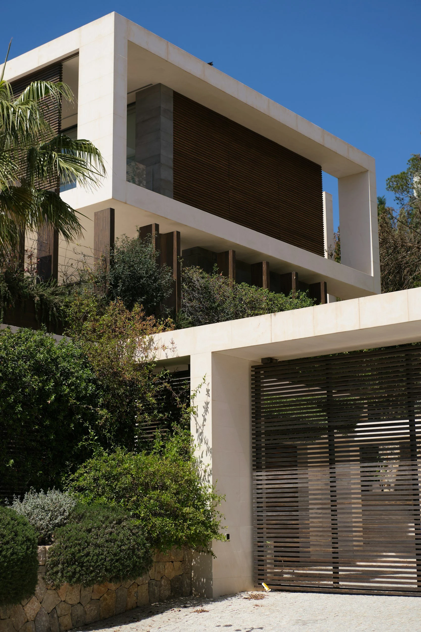 Modern house with white concrete and wooden accents, surrounded by green bushes and trees, under a clear blue sky.