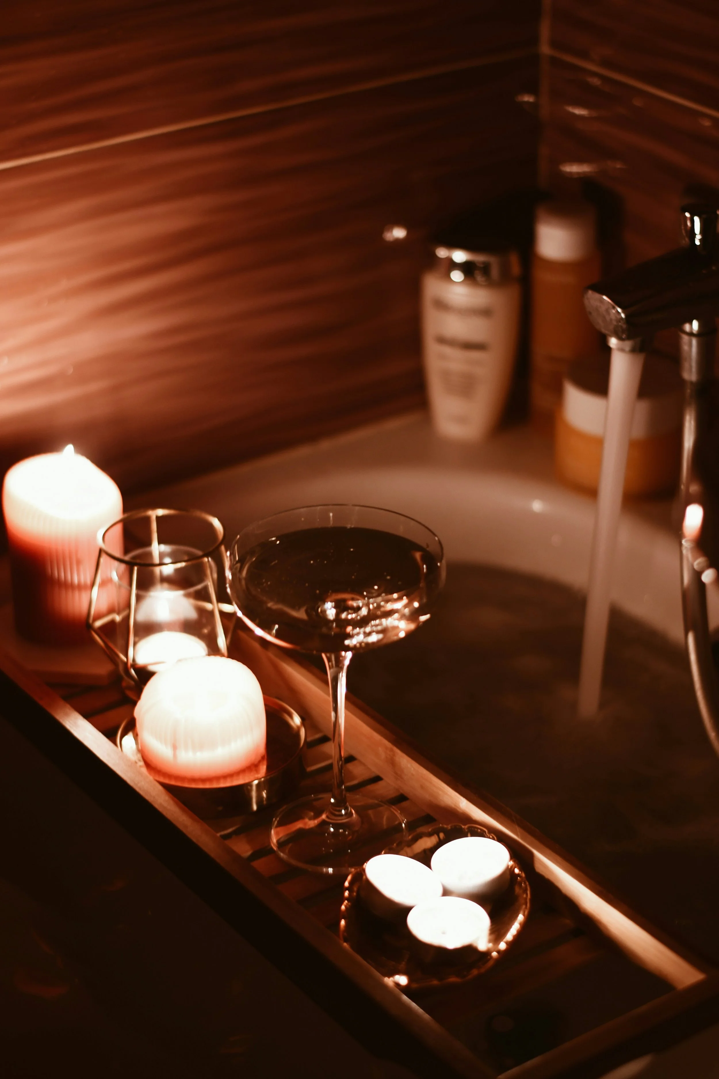 A dimly lit bathroom scene with candles, a cocktail glass, and a small tray on a wooden surface, nearby a bathtub with a faucet running.