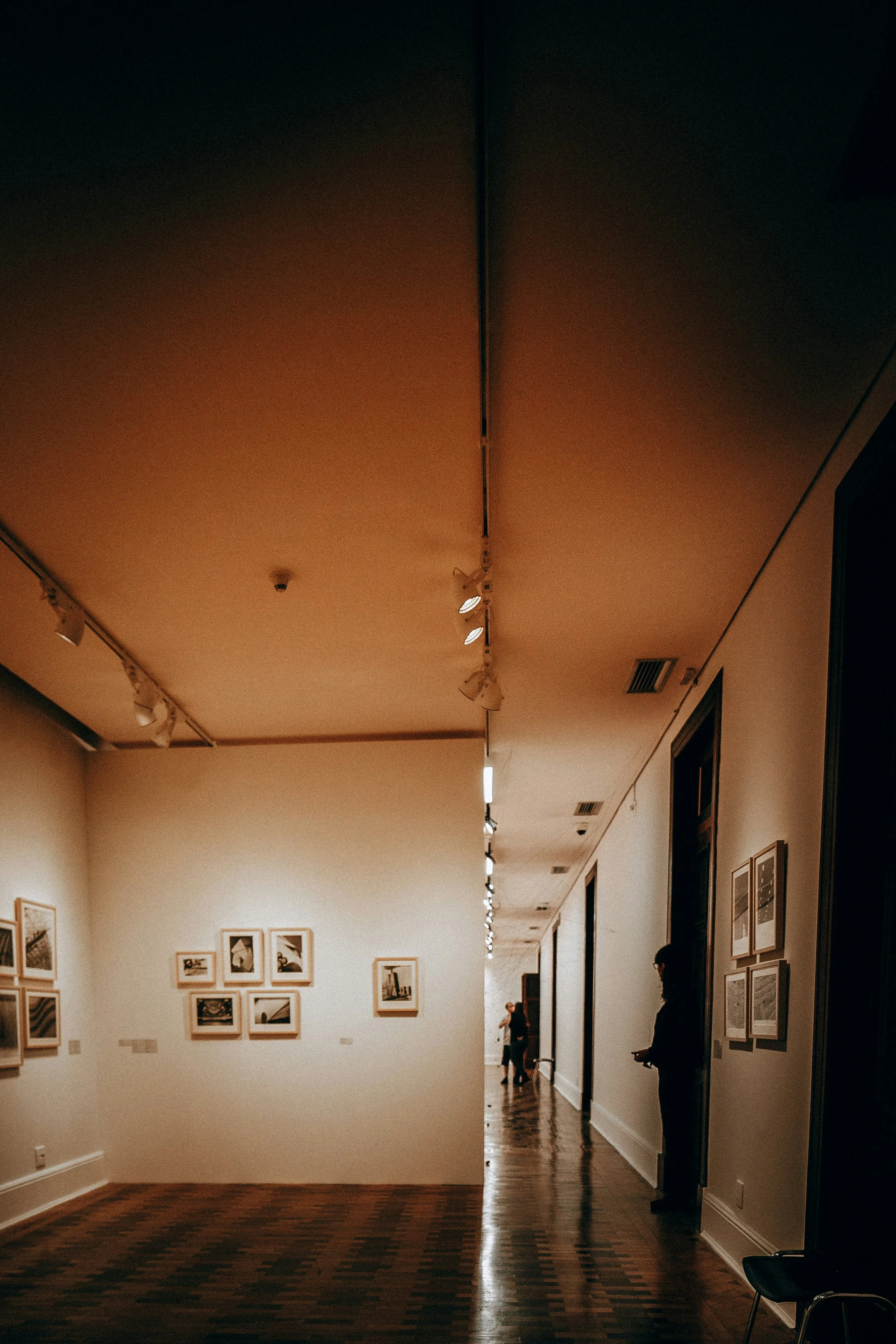 People observing framed photographs in an art gallery with wooden floors and minimal lighting.
