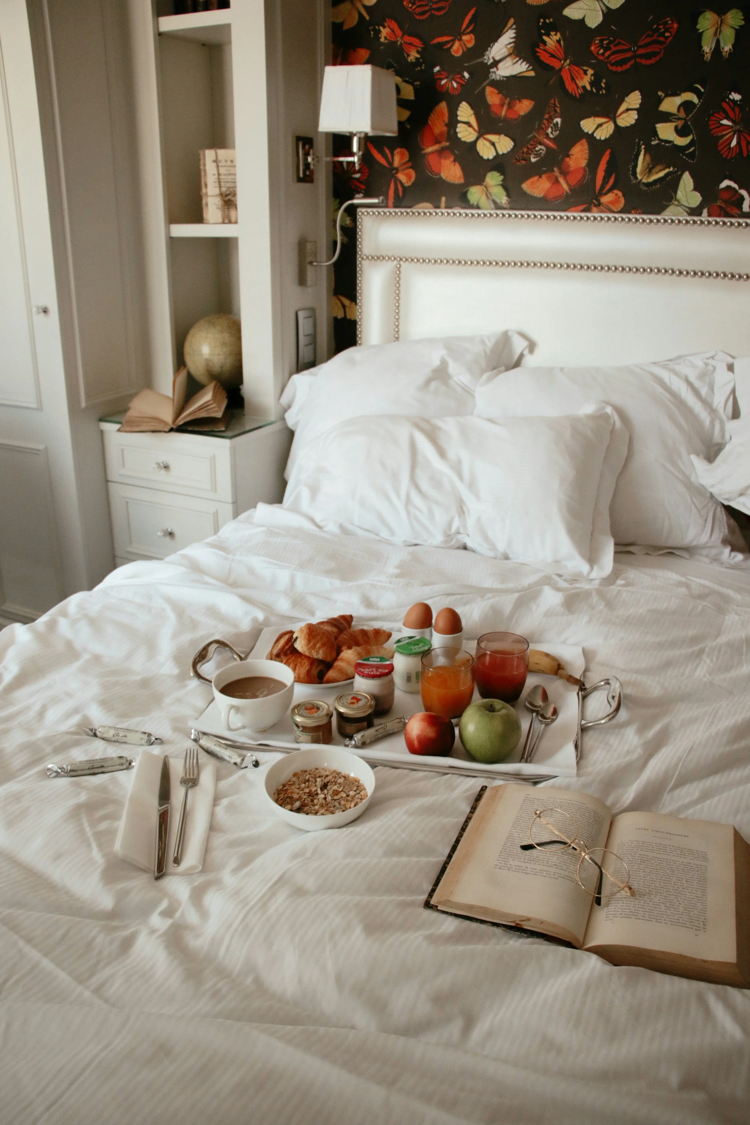 A breakfast tray with eggs, croissants, apples, juice, coffee, yogurt, and cereal on a bed. An open book and reading glasses are also on the bed. The background features a nightstand, a lamp, and a butterfly-patterned wallpaper.