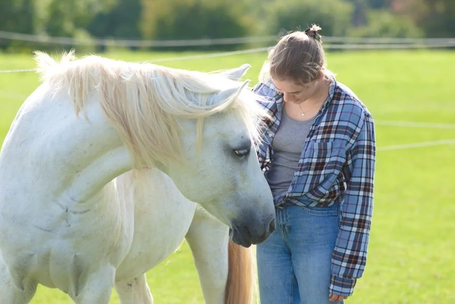 Eine Frau steht neben einem weißesn Pferd, welches den Kopf zu ihr dreht.