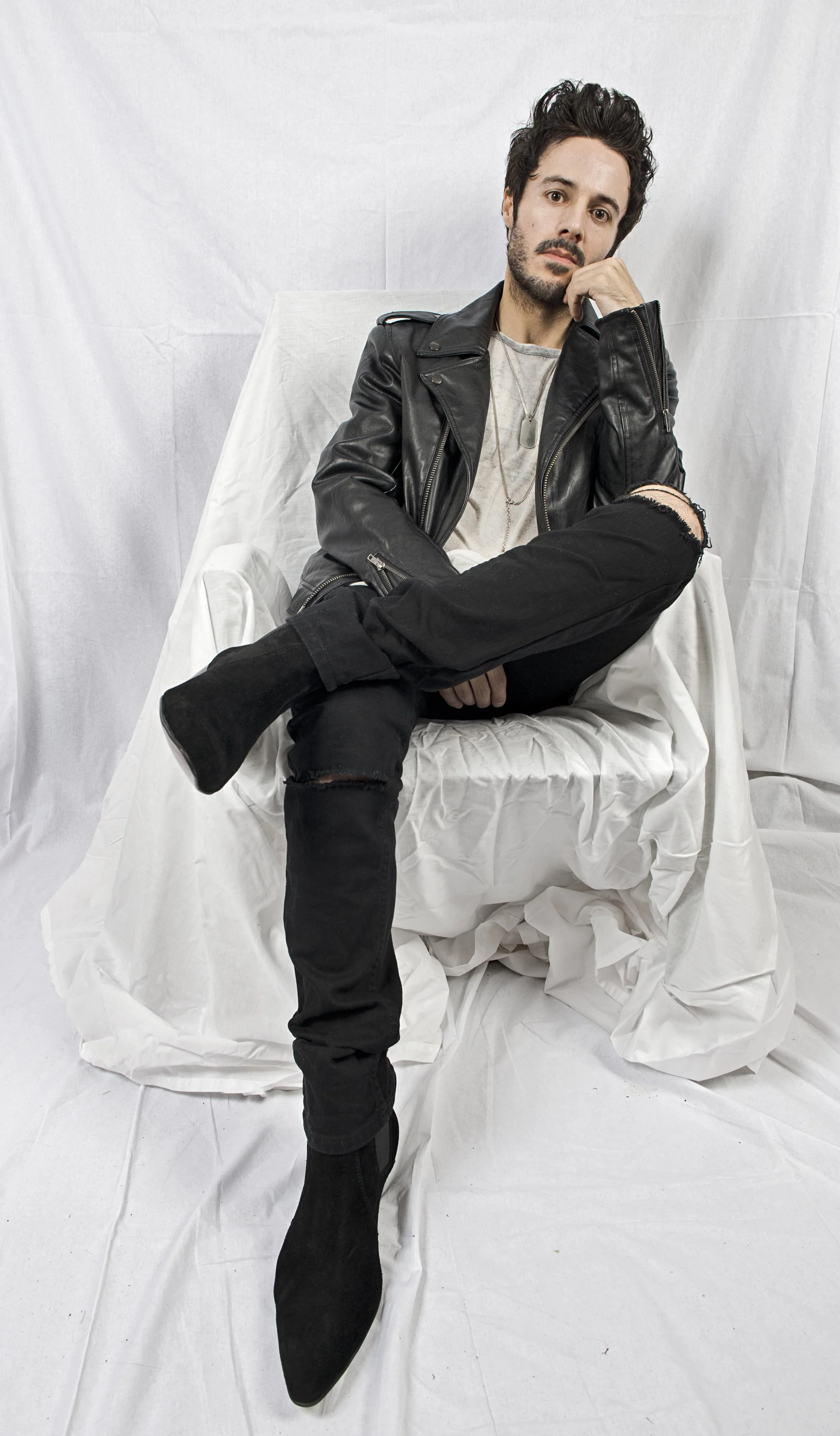James Kennedy, singer, author and frontman of the band 'Kyshera' posing in a white chair and wearing black boots and a leather jacket