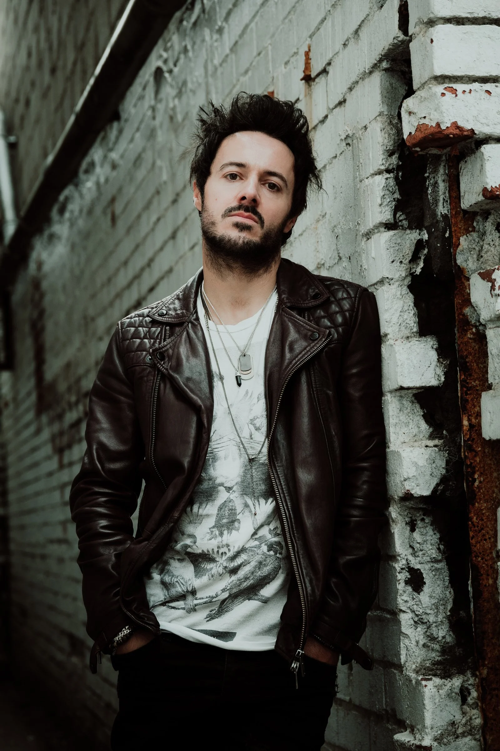 James Kennedy, the singer, author and podcaster from Wales leaning against a white wall and posing in a leather jacket for a photoshoot