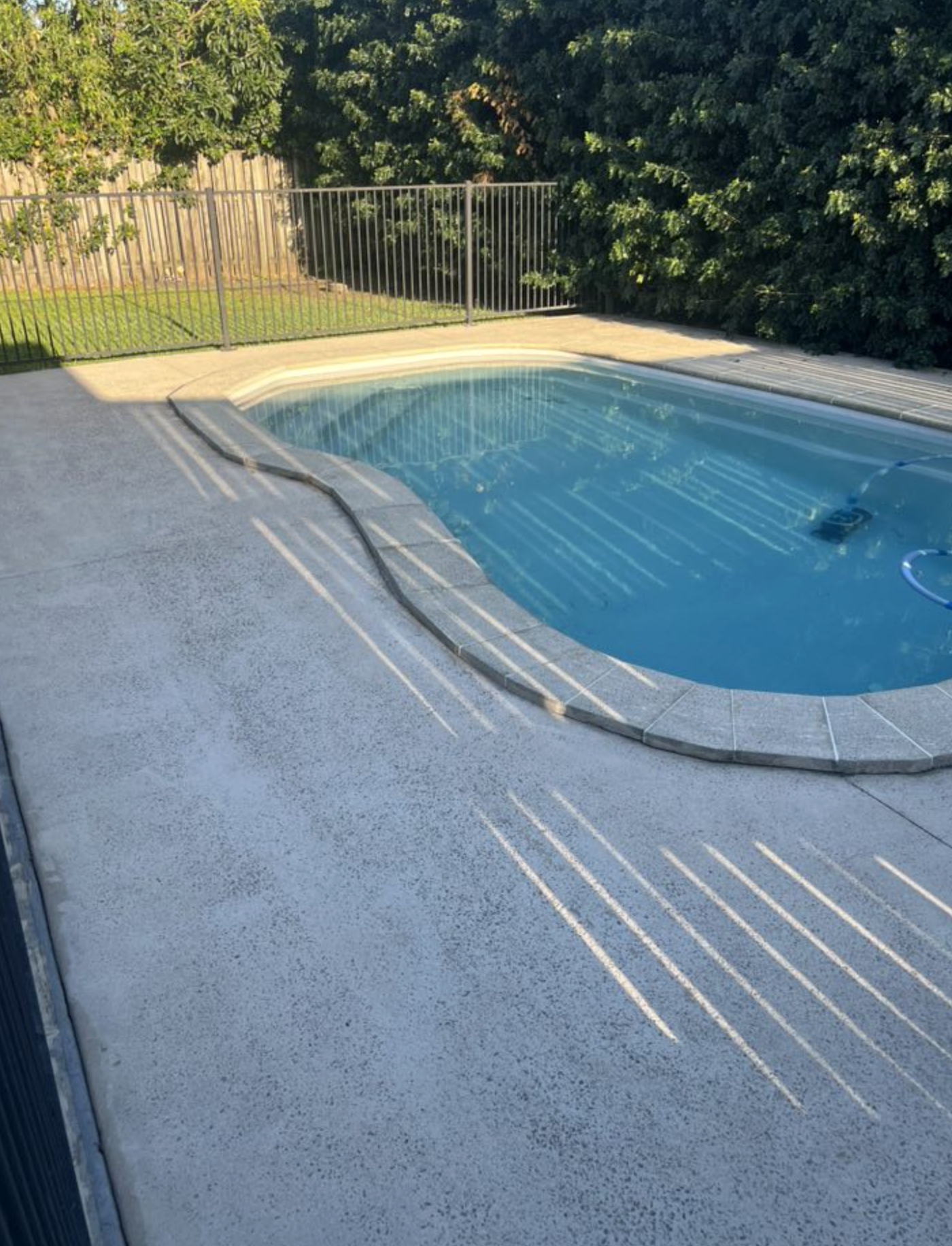 Small backyard swimming pool with blue water, surrounded by beige textured concrete patio and a black metal fence, with green bushes and trees in the background.