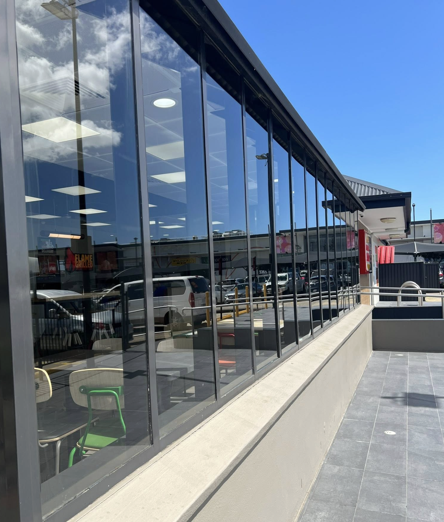 A view of the exterior of a restaurant with large glass windows reflecting the blue sky and clouds. Inside, tables and chairs are visible through the windows, and a parking lot with cars is seen outside.