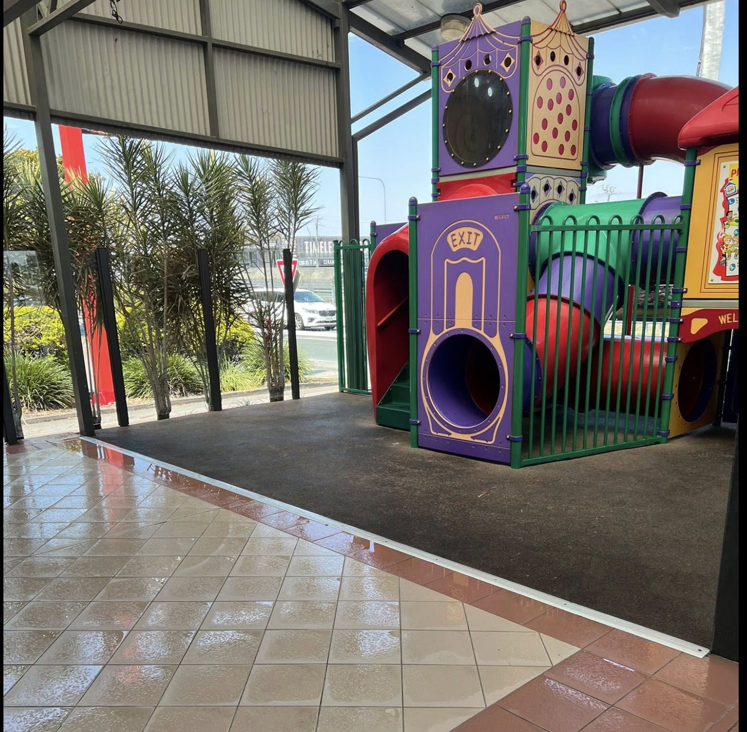 Colorful indoor playground structure with slides, tunnels, and an exit sign, adjacent to a glass wall with plants outside.