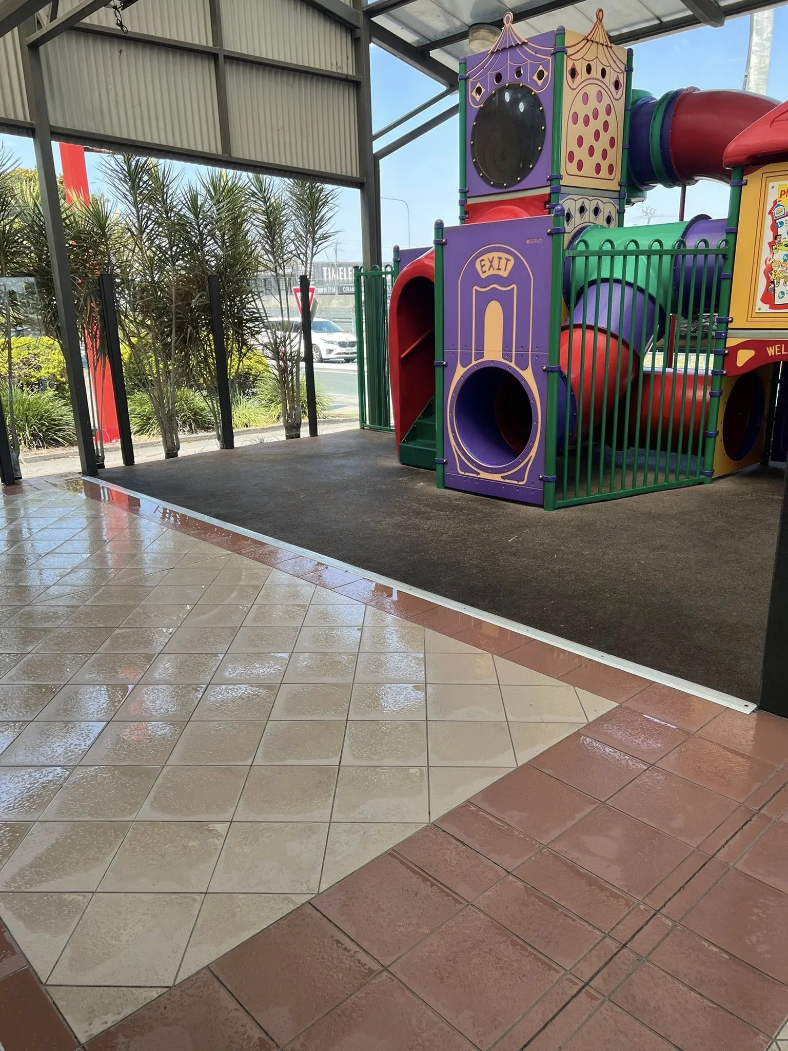 Colorful indoor children's play structure with slides and tunnels, labeled EXIT, in a shopping mall or similar environment, next to tiled floor and large glass windows.