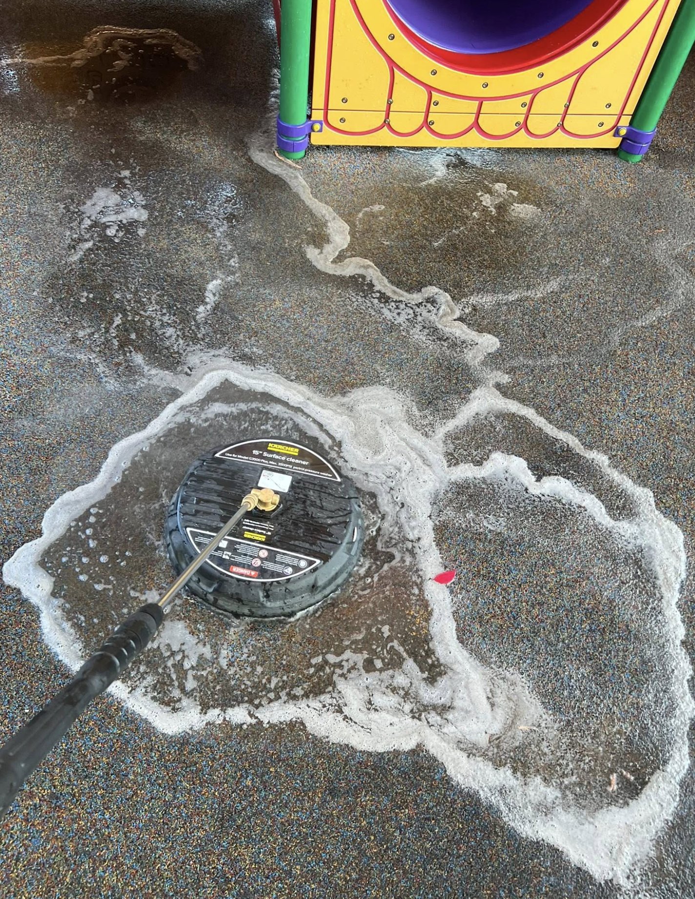 A floor cleaning machine is being used to clean a wet, textured carpeted floor. The floor is covered with soap suds and water, with foam around the machine. Part of a colorful plastic play structure with yellow, green, purple, and red colors is visib