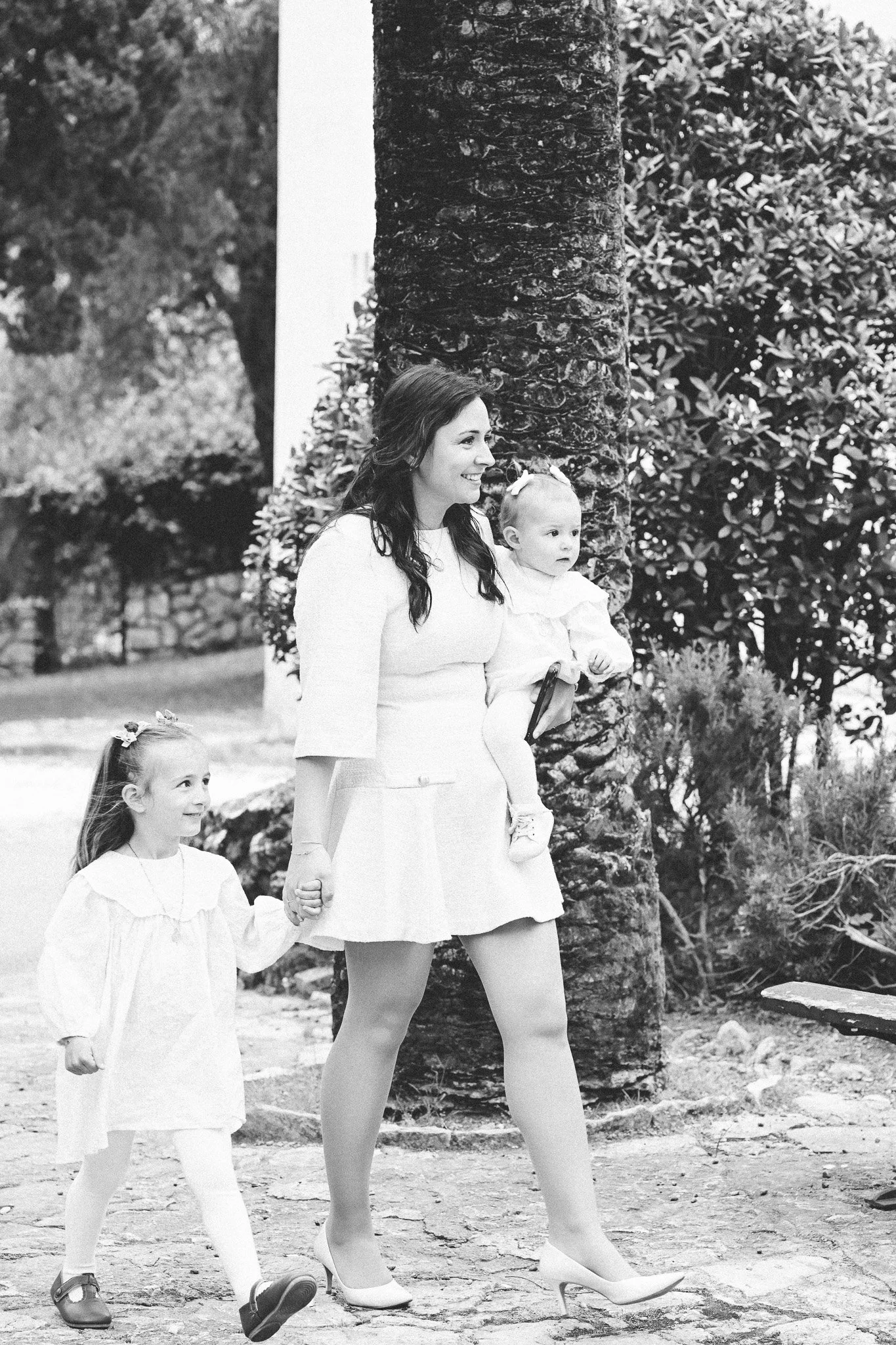 Une femme avec deux jeunes filles, marchant dans un parc. La femme porte une jupe et une paire de talons, avec les cheveux longs et ondulés. La plus jeune fille tient la main de la femme et porte une robe blanche, tandis que l'autre fille marche à cô