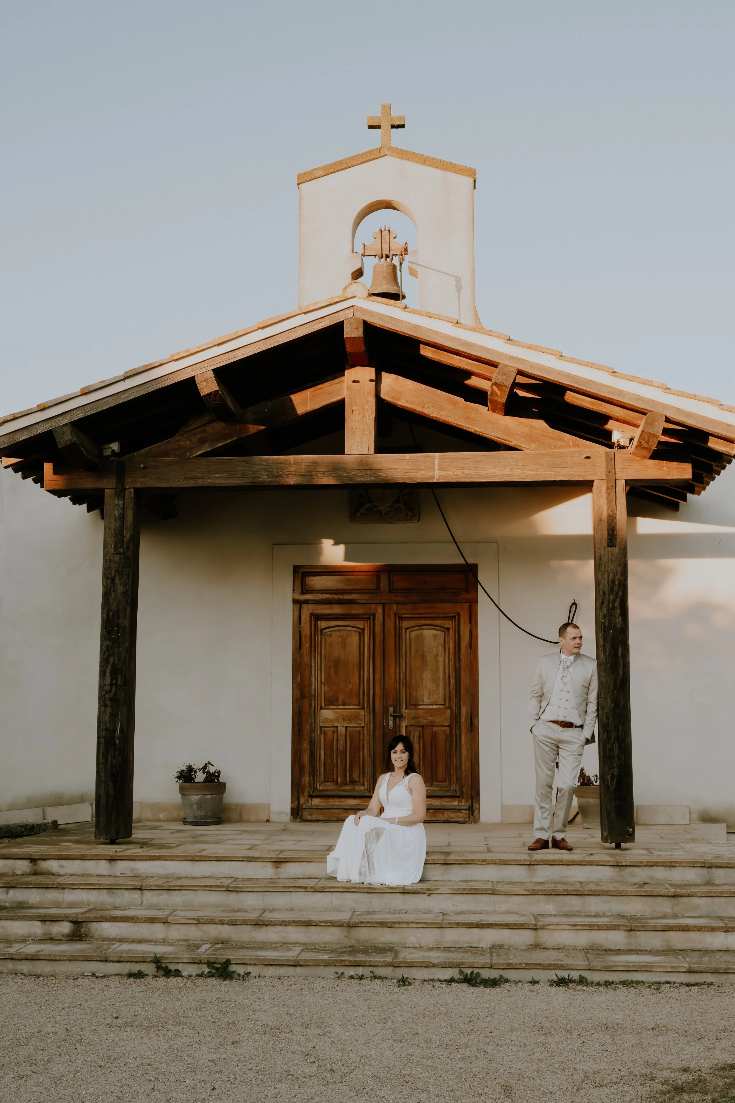 Une femme en robe blanche assise sur les escaliers devant une porte en bois, un homme en costume blanc setenant contre un poteau en bois à côté d'elle, à l'extérieur d'une église avec un petit clocher et une croix au sommet, sous un ciel clair