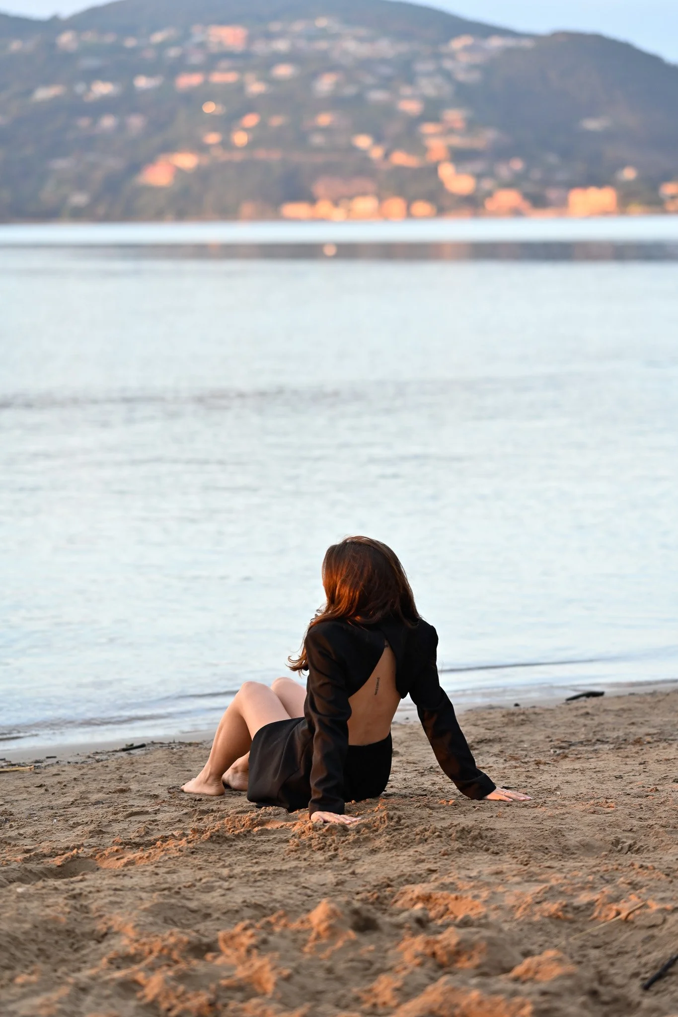 Une femme avec des cheveux bruns, portant une veste noire et une jupe noire, assise sur la plage de sable, regardant vers la mer, avec une montagne en arrière-plan.