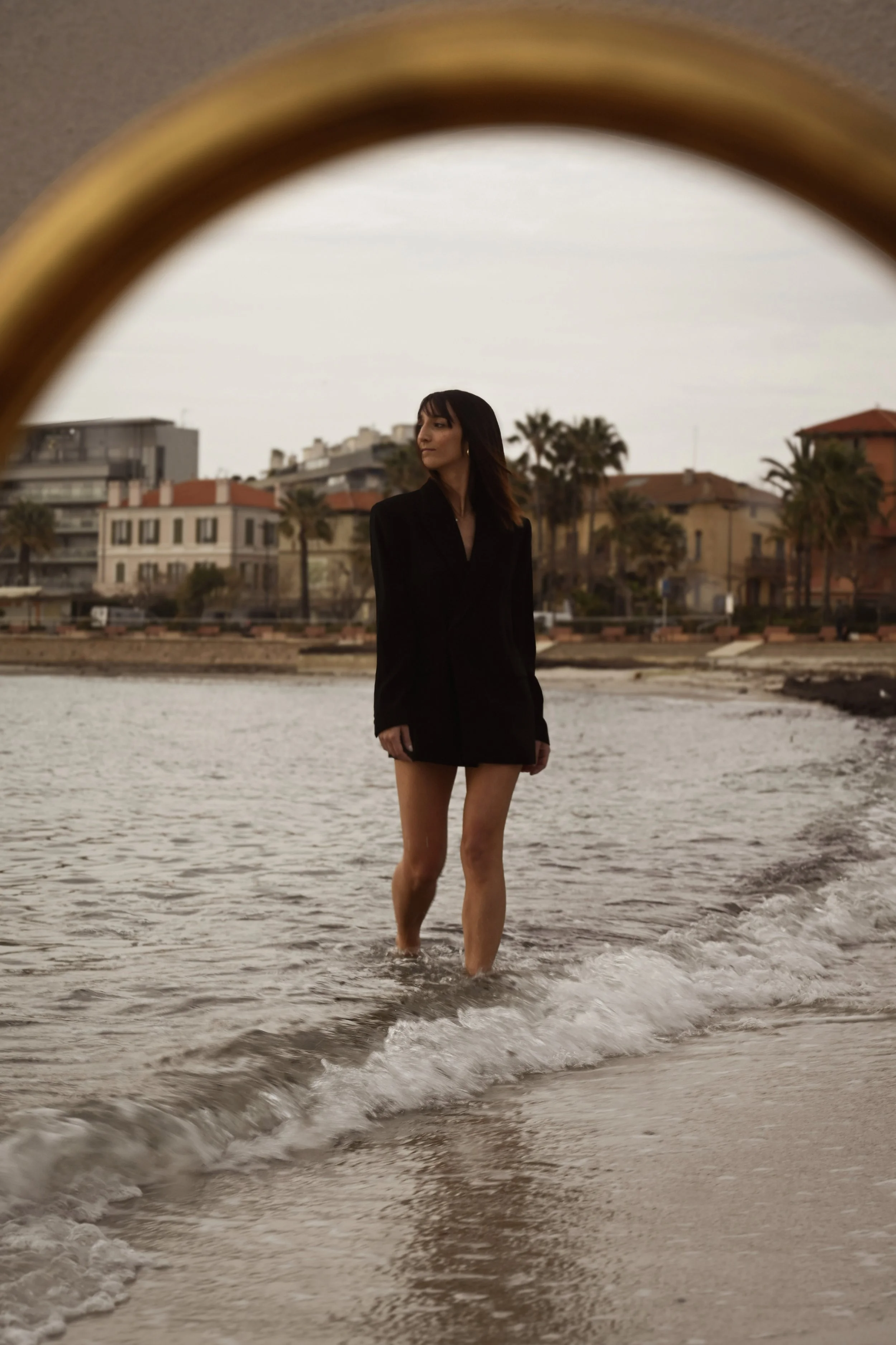 Une femme portant une veste noire et un short marche dans l'eau de la mer, avec des maisons et des palmiers au fond, sous un ciel nuageux.