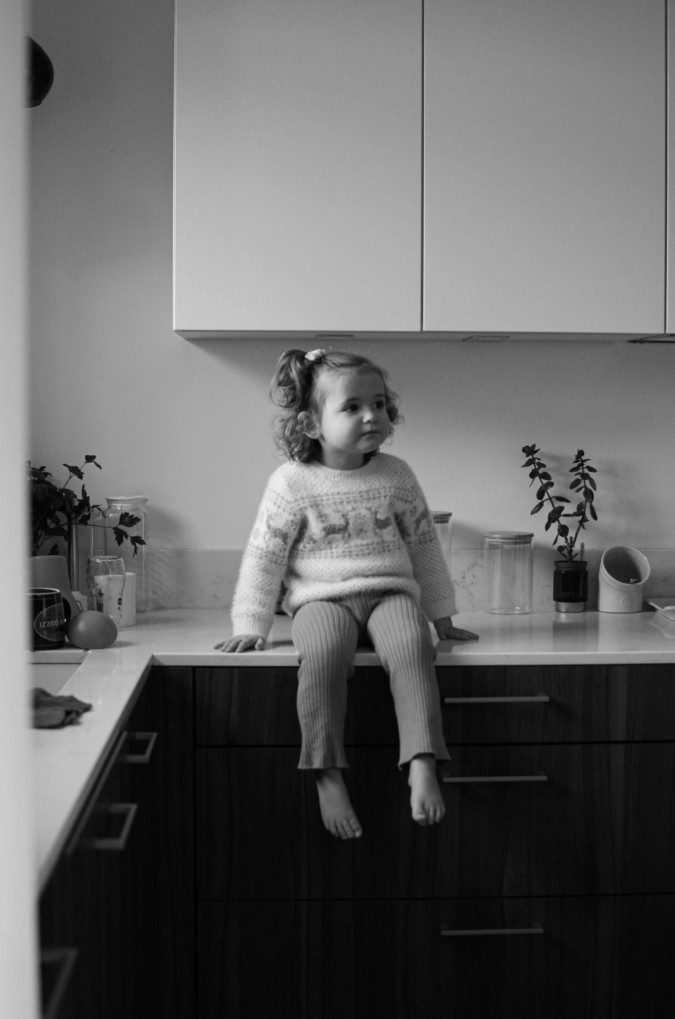 Une petite fille assise sur un comptoir de cuisine, portant un pull à motifs et des collants rayés. Elle regarde vers la droite, avec des plantes en pot et des bouteilles en verre en arrière-plan.