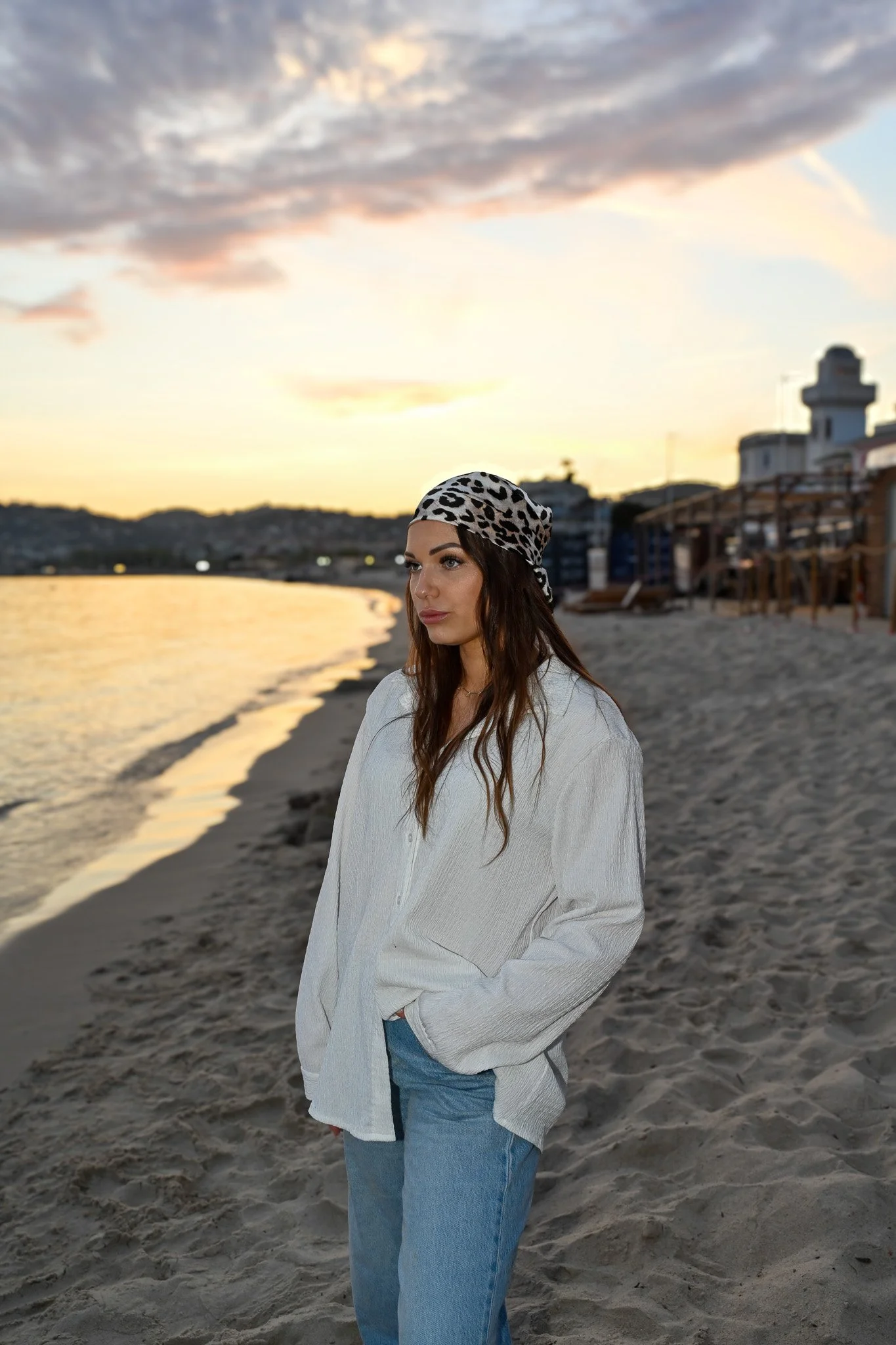 Une femme debout sur une plage au coucher du soleil, portant une veste blanche, un jean bleu, et un bandana léopard sur la tête, avec des bâtiments en arrière-plan.