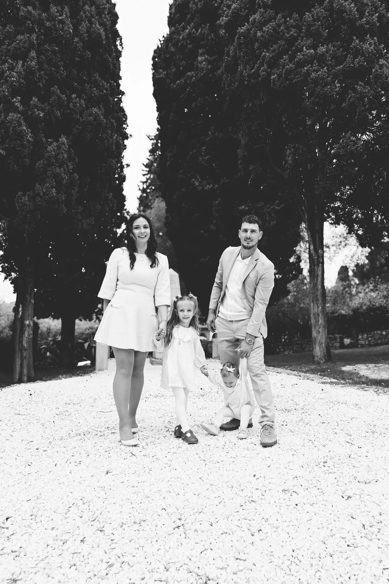 Une famille se promène dans un parc avec des arbres en arrière-plan, tous souriants et habillés de manière élégante, la maman en robe blanche, le papa en costume clair, deux jeunes filles en robes blanches, une d'elles portant une chaussure sur le so