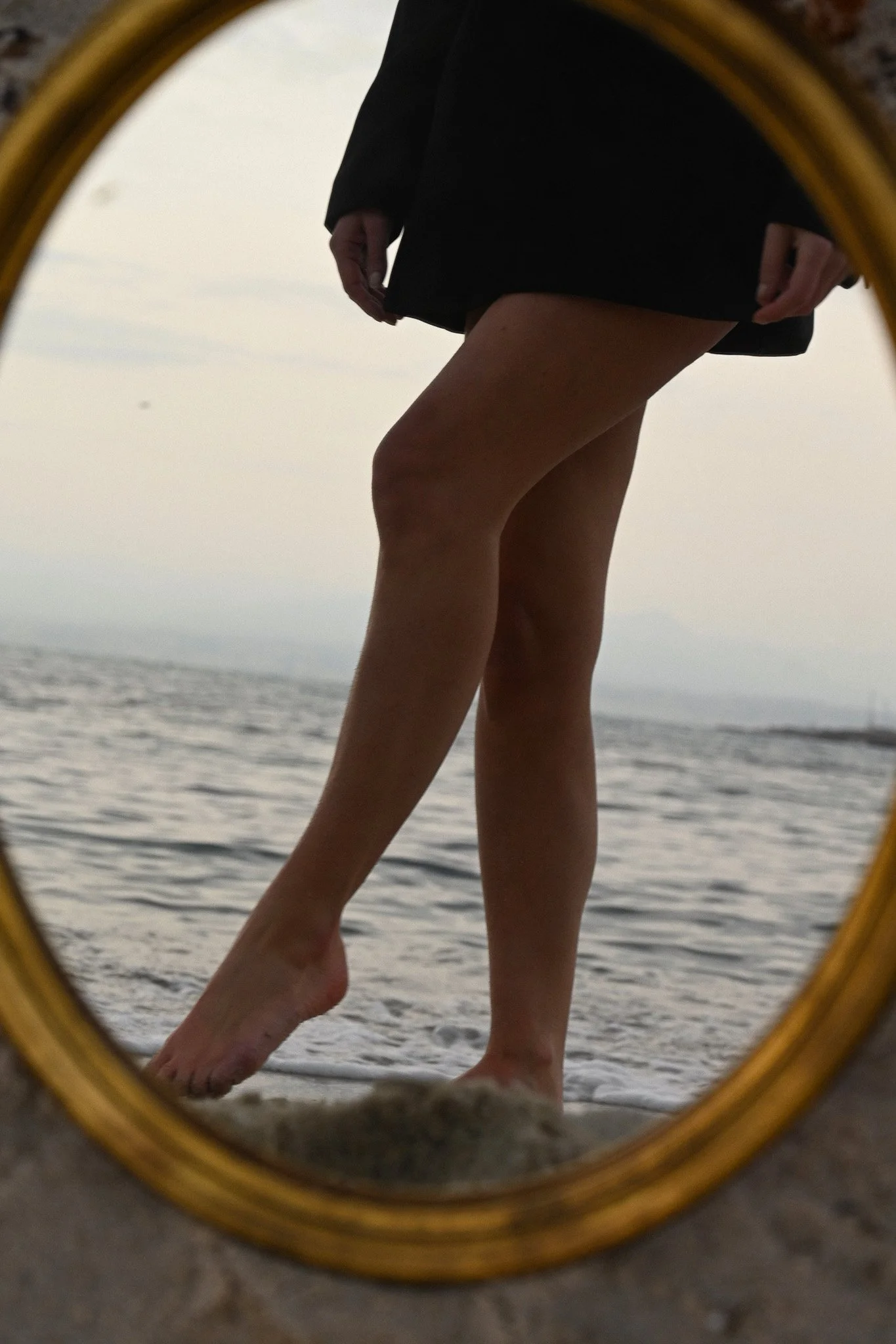 Reflet d'une personne en maillot noir dans un miroir rond placé sur le sable près de la mer, avec le ciel nuageux en arrière-plan.