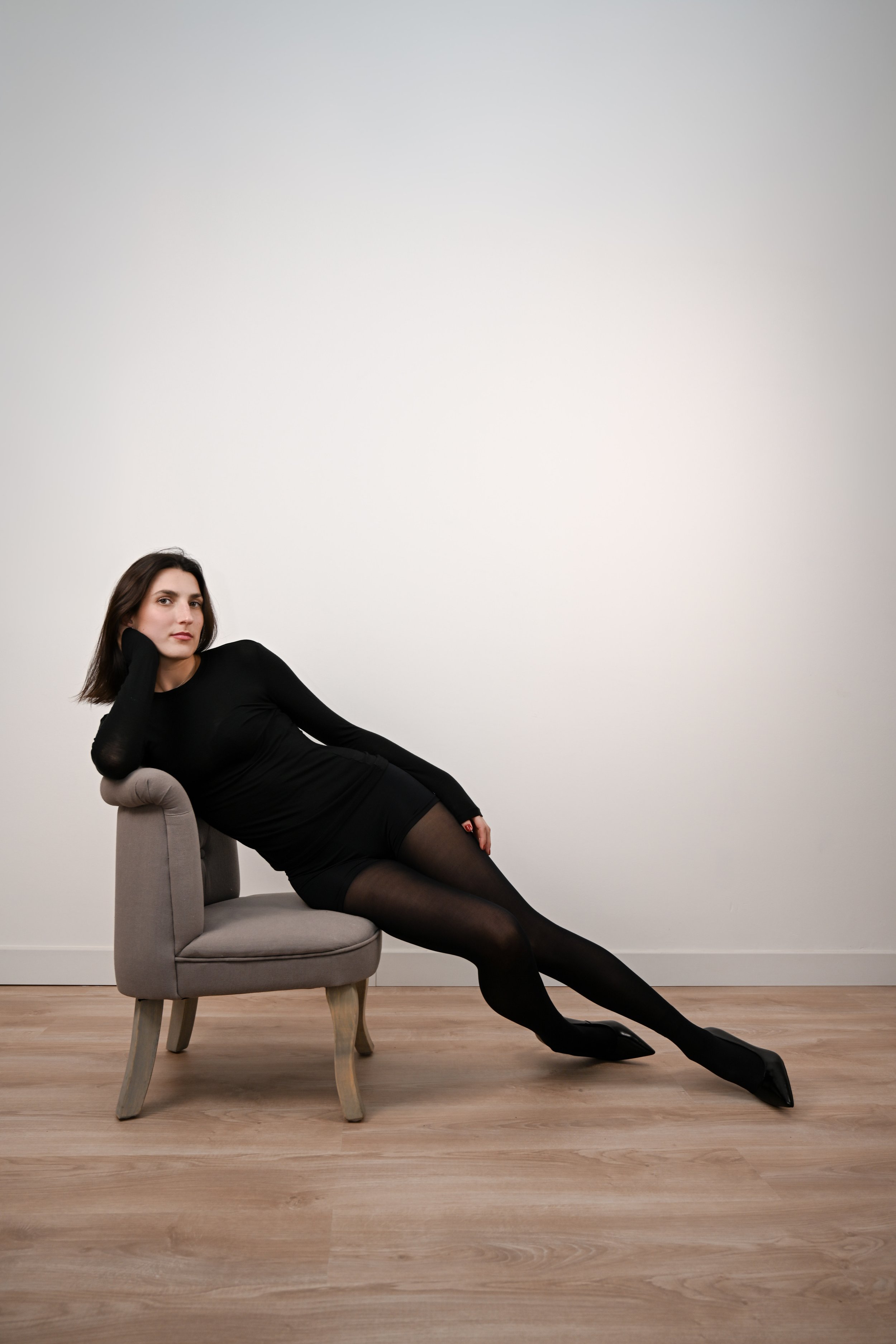 Une femme habillée en noir, assise dans un fauteuil, repose la tête sur sa main, position allongée, dans une pièce avec un mur blanc et un sol en bois.