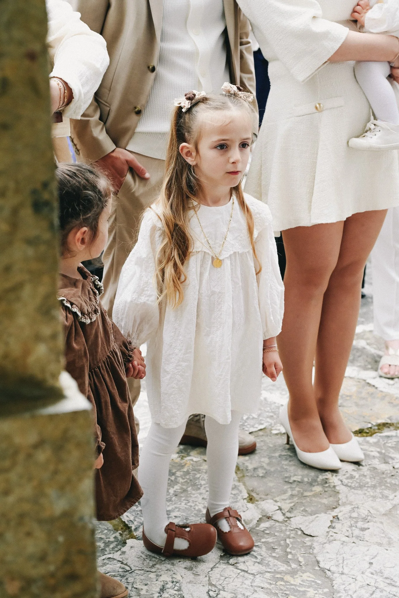 Jeune fille avec une robe blanche, collier doré et cheveux avec des attaches en tissu, en groupe lors d'un événement en plein air.