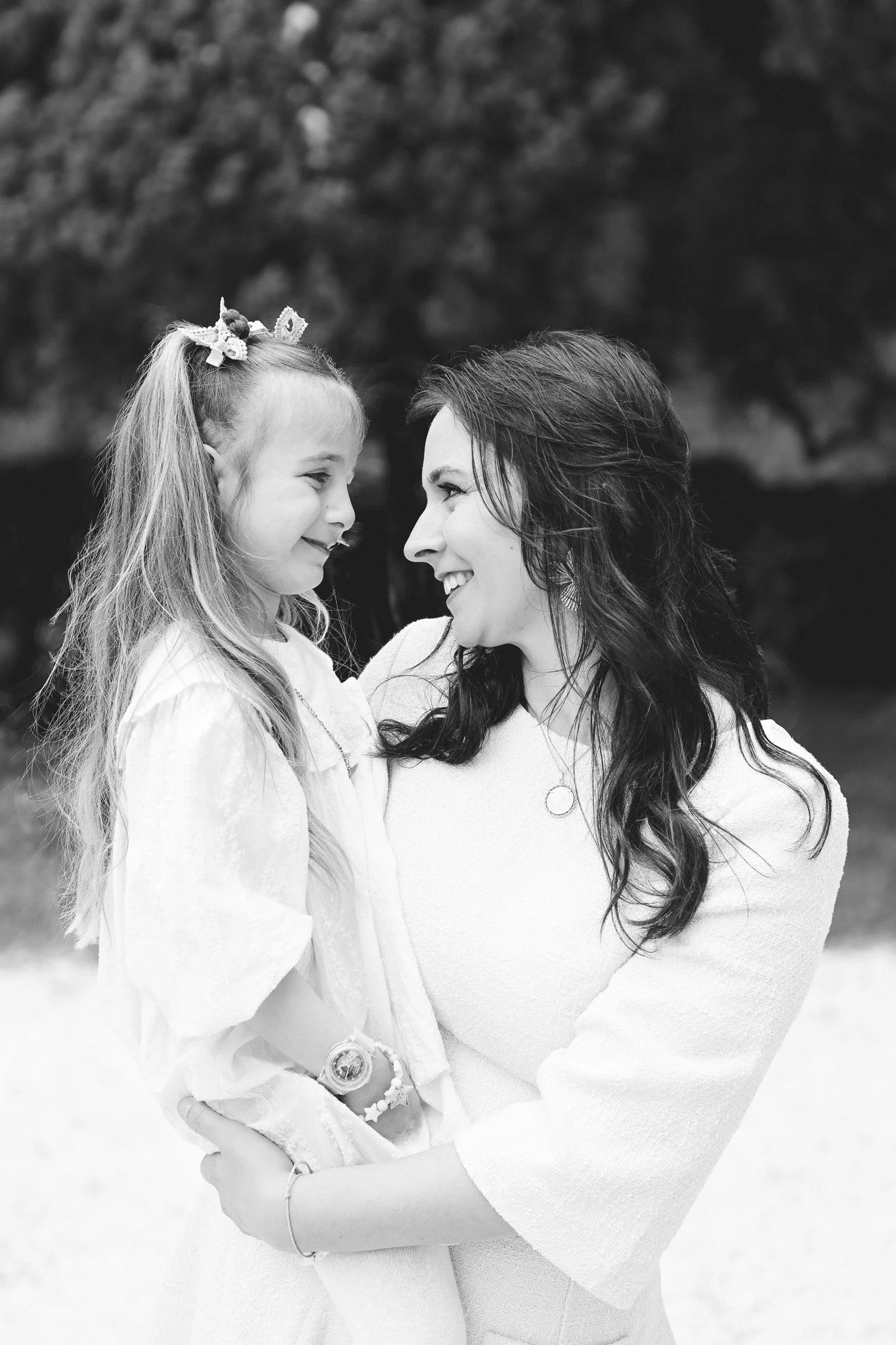 Une femme et une jeune fille souriantes se regardant tendrement, en noir et blanc, dehors avec des arbres flous en arrière-plan.
