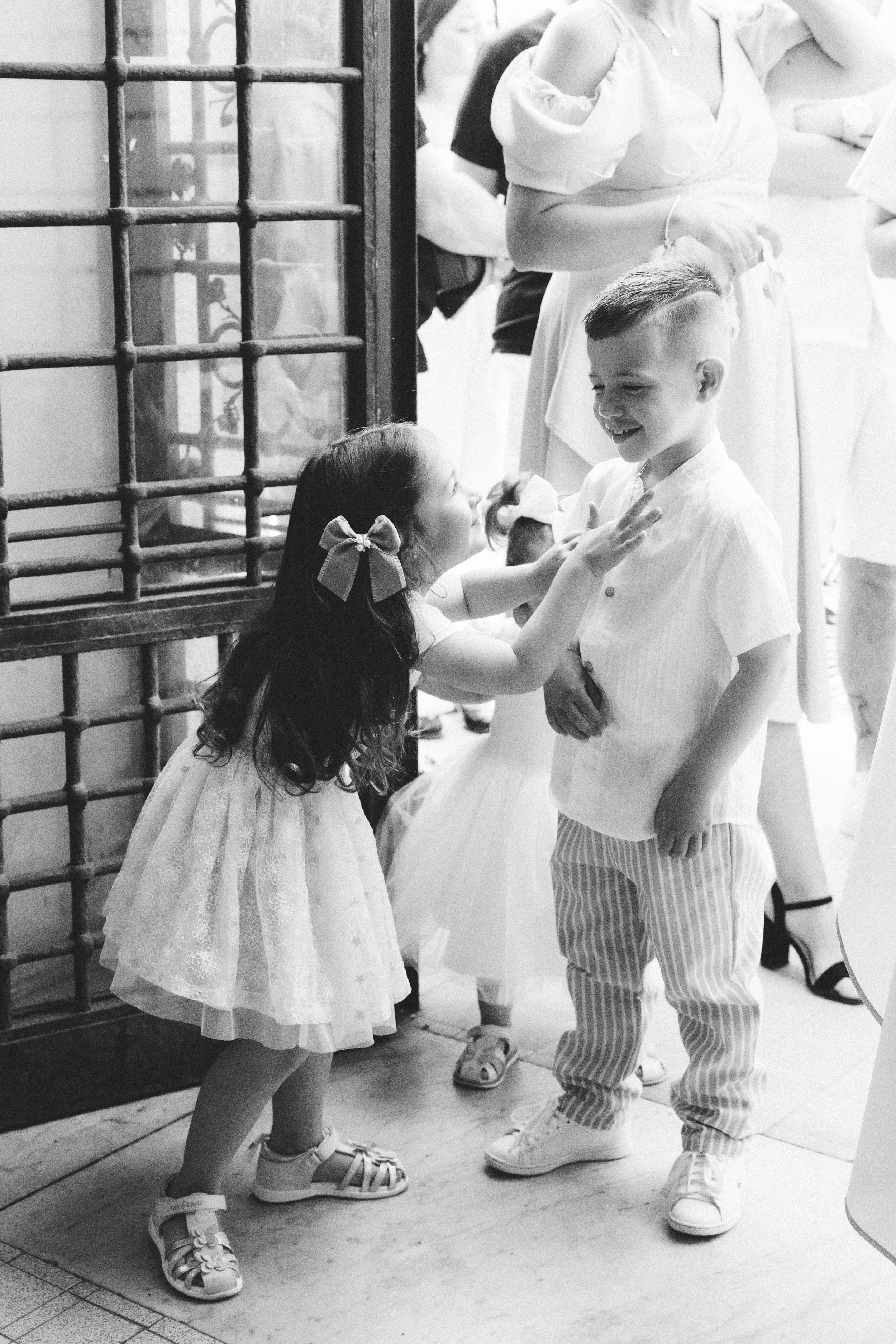 Une petite fille avec une robe blanche et un nœud dans les cheveux parlant à un garçon avec une chemise blanche et un pantalon rayé. Autres enfants en arrière-plan, dans un environnement intérieur.