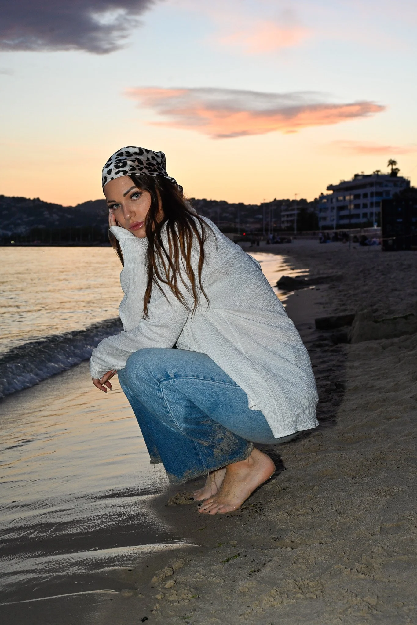 Jeune femme assise sur le rivage de la mer au coucher du soleil, portant un bandana à motifs léopard, une chemise blanche et un jean, avec des bâtiments et des arbres en arrière-plan.