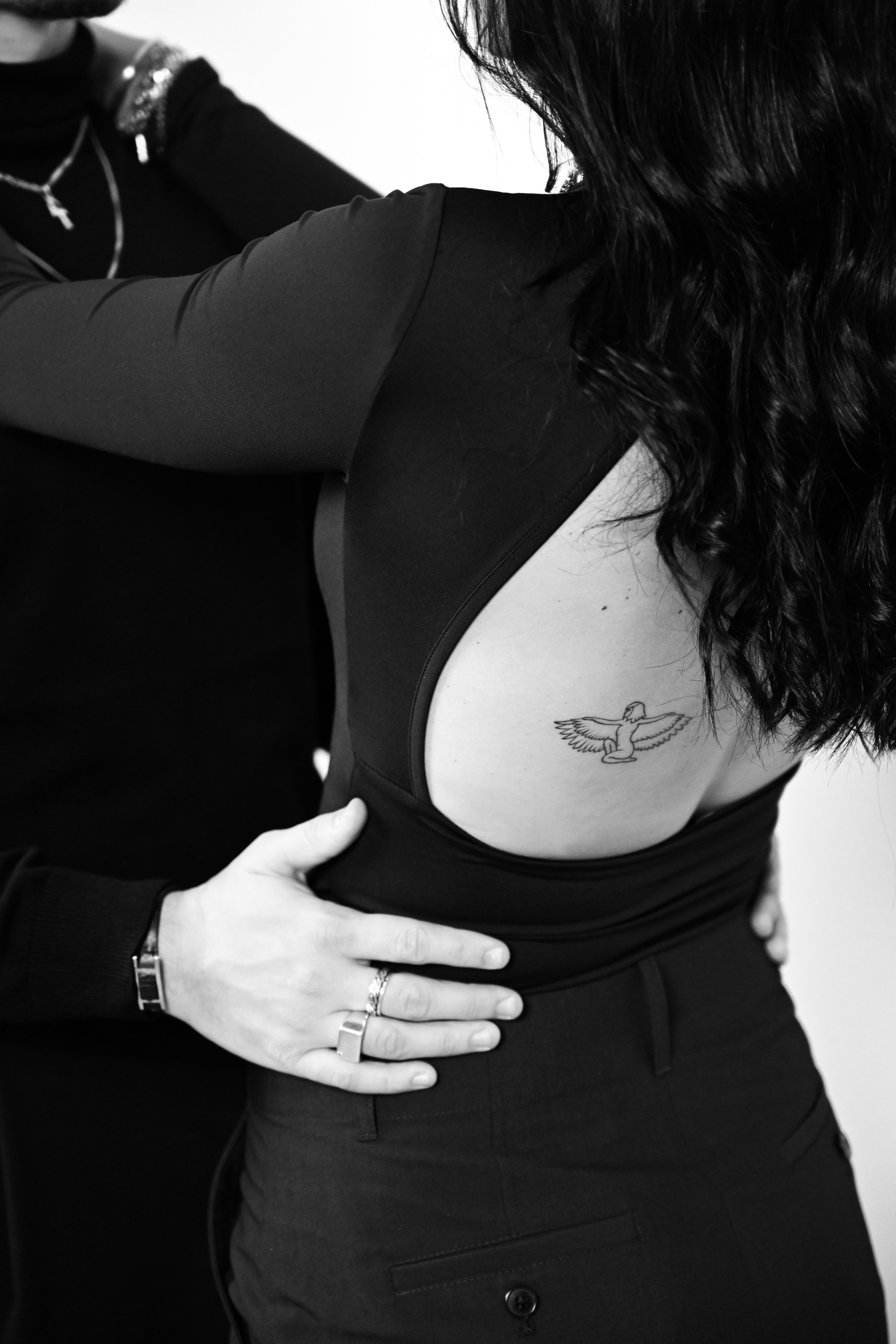 Une femme avec des cheveux longs portant un haut noir laissant apparaître un tatouage d'une femme ailée sur la côte, tenant une autre personne dans une embrassade.