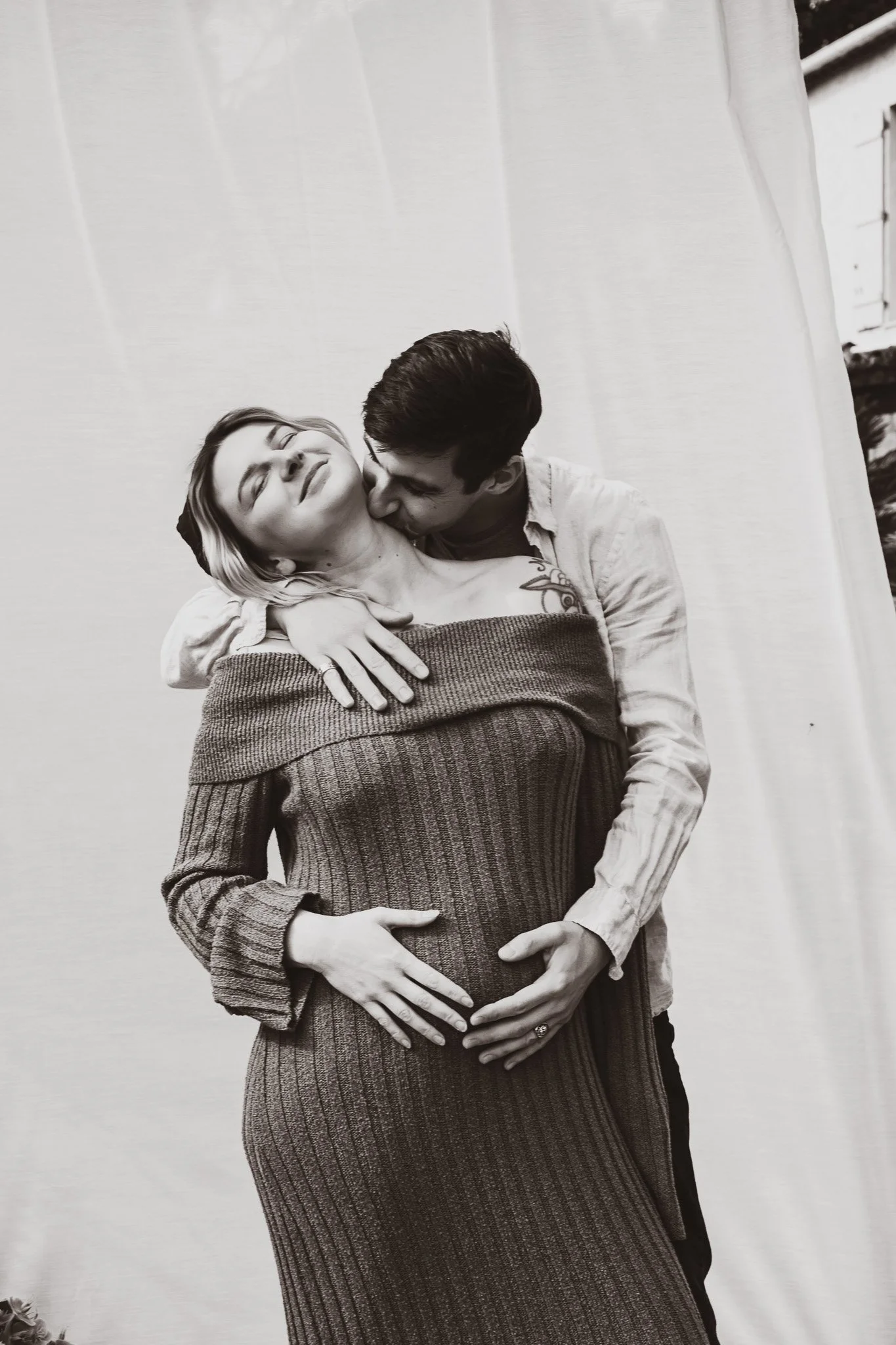 Un couple s'embrasse tendrement, la femme est enceinte, elle porte un pull à rayures et la femme a un tatouage sur le bras.