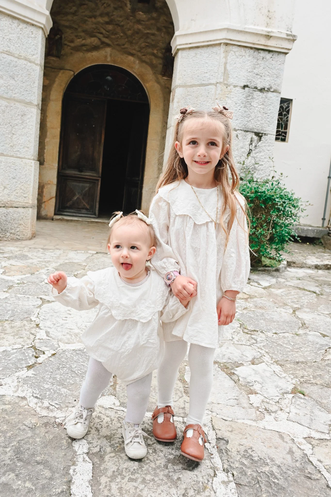Deux jeunes filles en tenues blanches posant dehors devant une porte en pierre. La plus grande a les cheveux coiffés avec des rubans et porte une robe avec un collier. La plus petite a des nœuds dans les cheveux, une chemise et un pantalon, avec une 