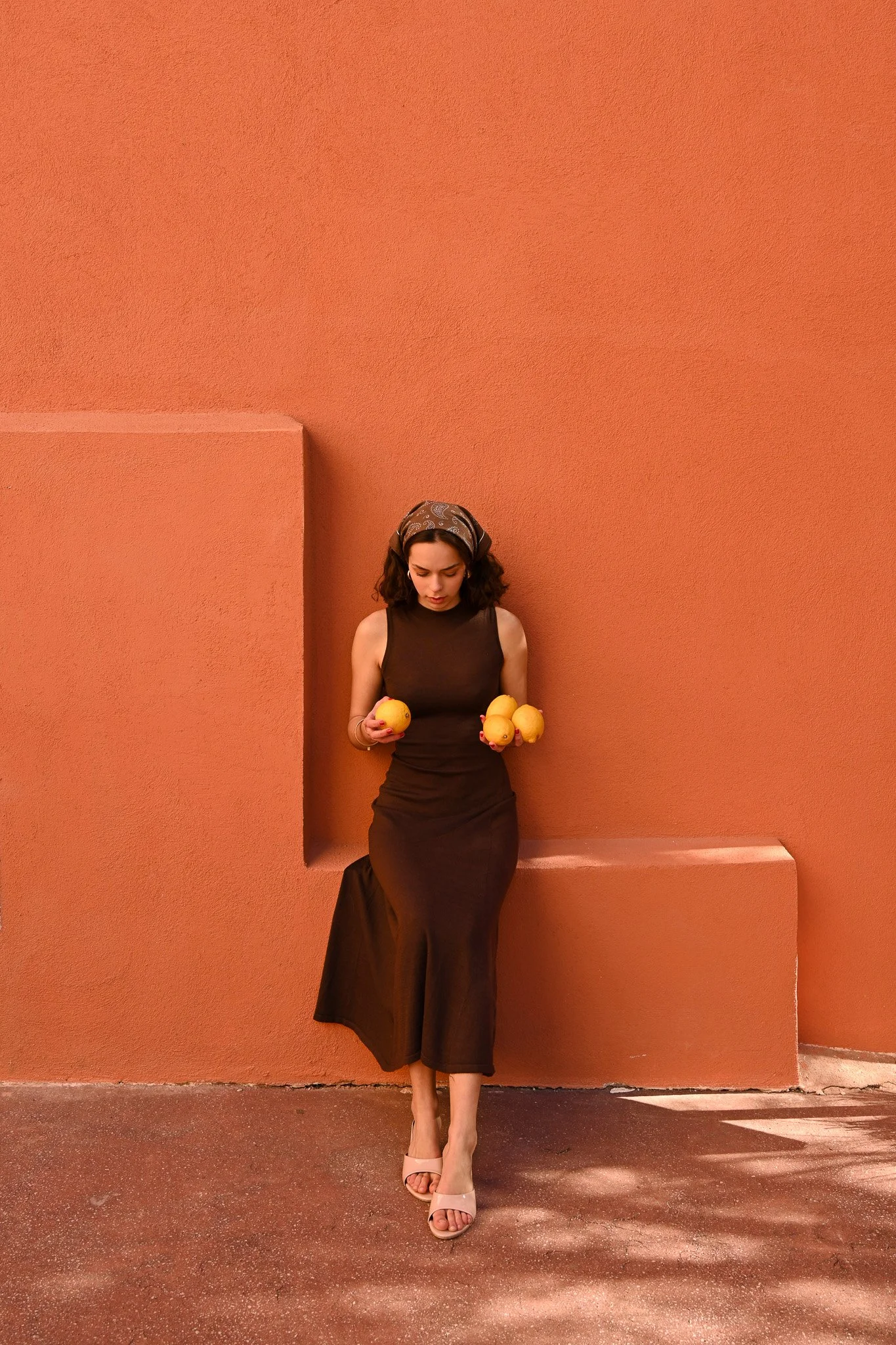 Une femme en robe noire, portant un foulard à motifs, tient quatre agrumes dans ses mains, debout devant un mur orange avec des formes géométriques.