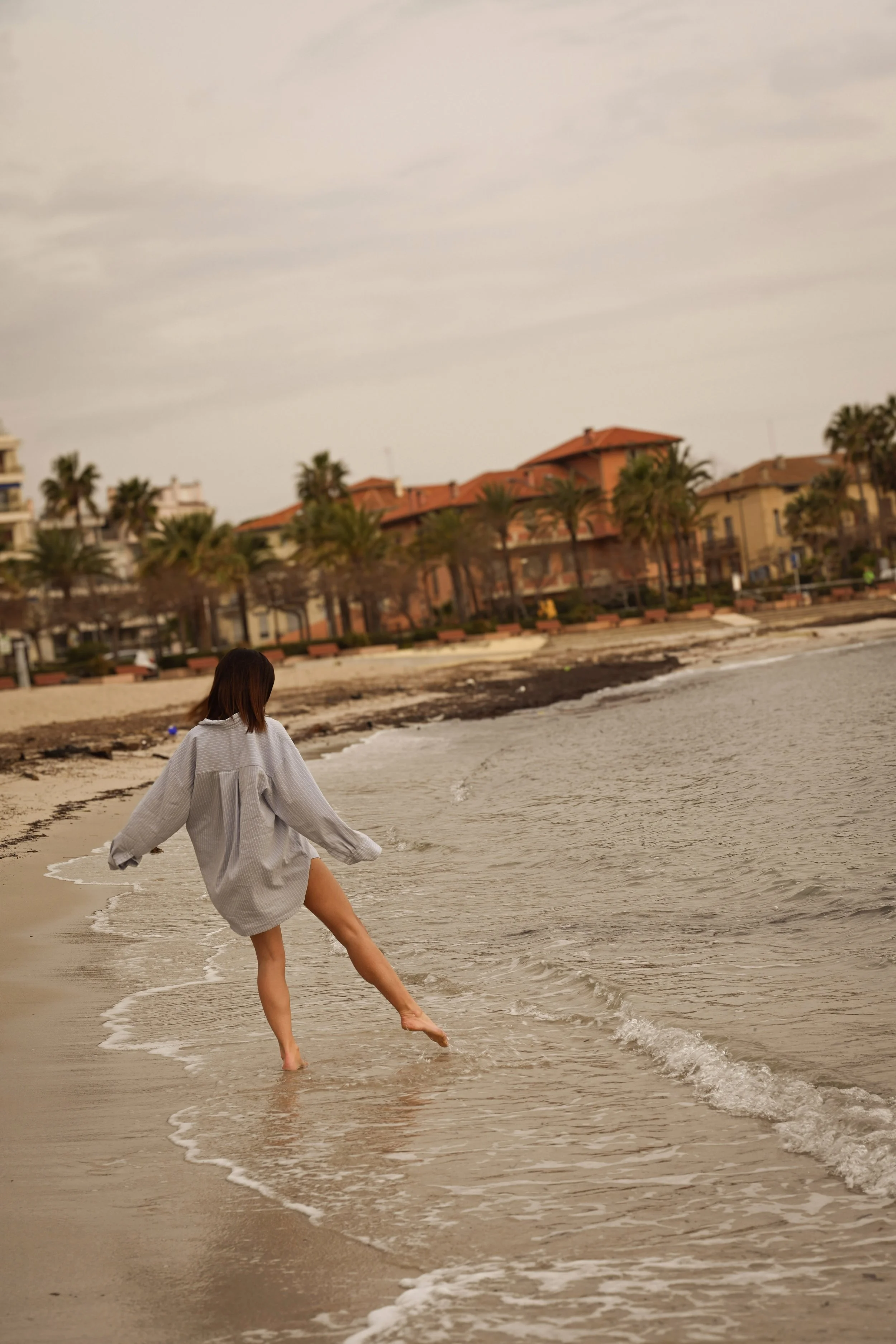Femme debout dans l'eau à la plage, portant une robe gris clair, avec des bâtiments et palmiers en arrière-plan, ciel nuageux au crépuscule.
