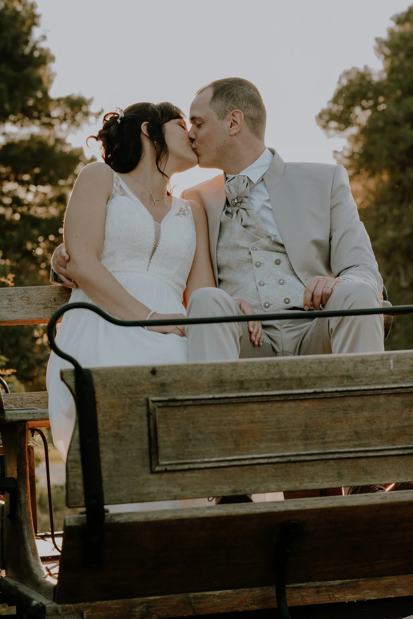 Un couple en mariage s'embrasse sur un banc en plein air pendant le coucher du soleil, avec des arbres en arrière-plan.