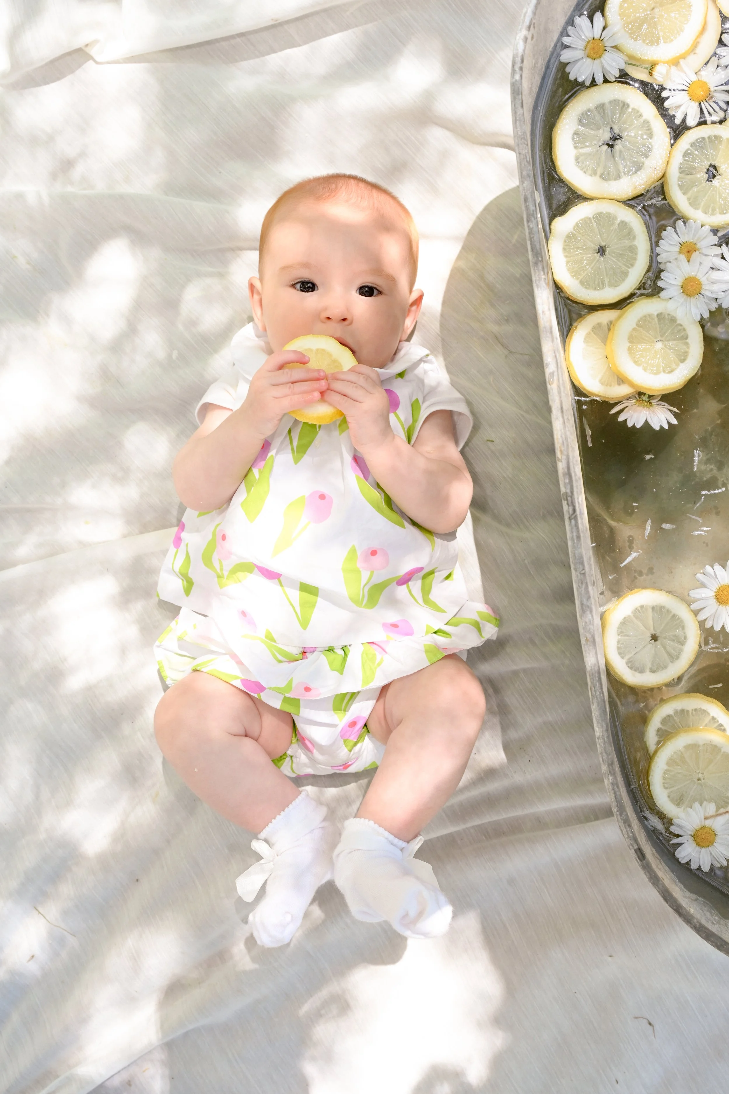 Un bébé allongé sur un lit en métal, tenant et mordant un morceau de citron, portant un pyjama à motifs de flamants roses et de feuilles, avec un plateau de tranches de citron dans de l'eau aromatisée avec des fleurs blanches.