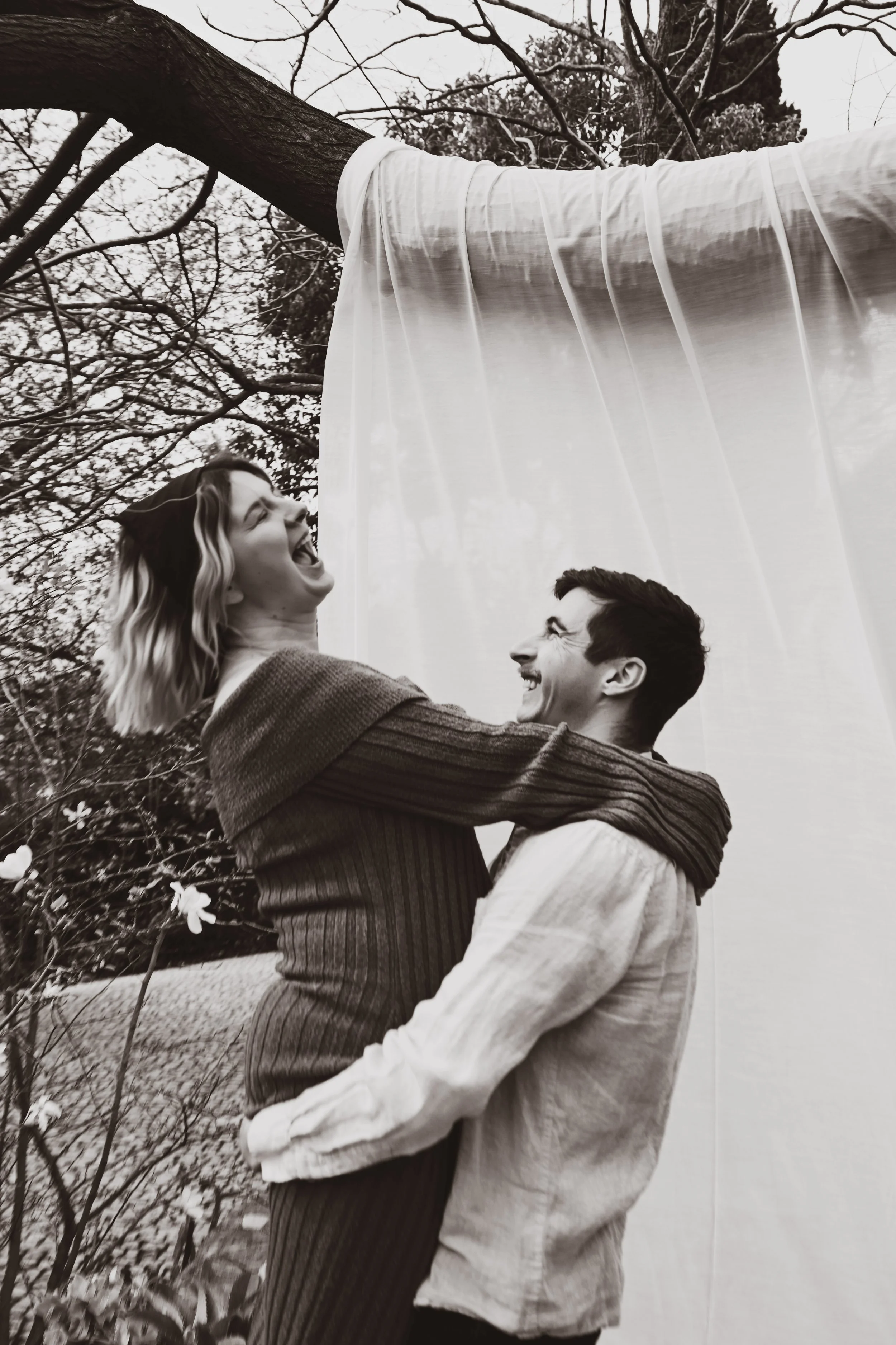 Un couple souriant, en plein air, avec un arbre en arrière-plan, l'homme porte un pull rayé et la femme une veste claire, ils semblent heureux et complices.