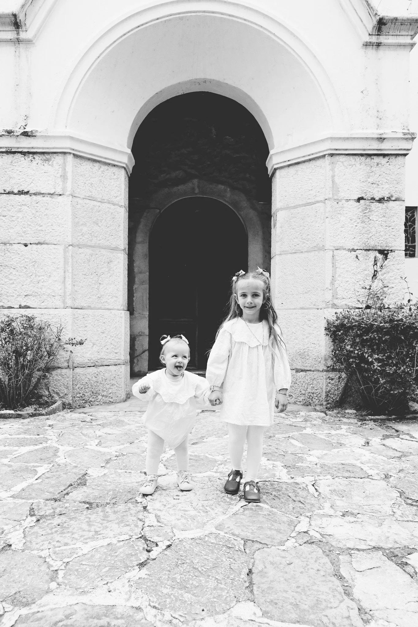 Deux jeunes filles souriantes marchant sur une rue pavée en face d'une vieille bâtisse en pierre blanche avec une arche. La plus âgée tient la main de la plus jeune, toutes deux portent une robe blanche et ont les cheveux attachés.