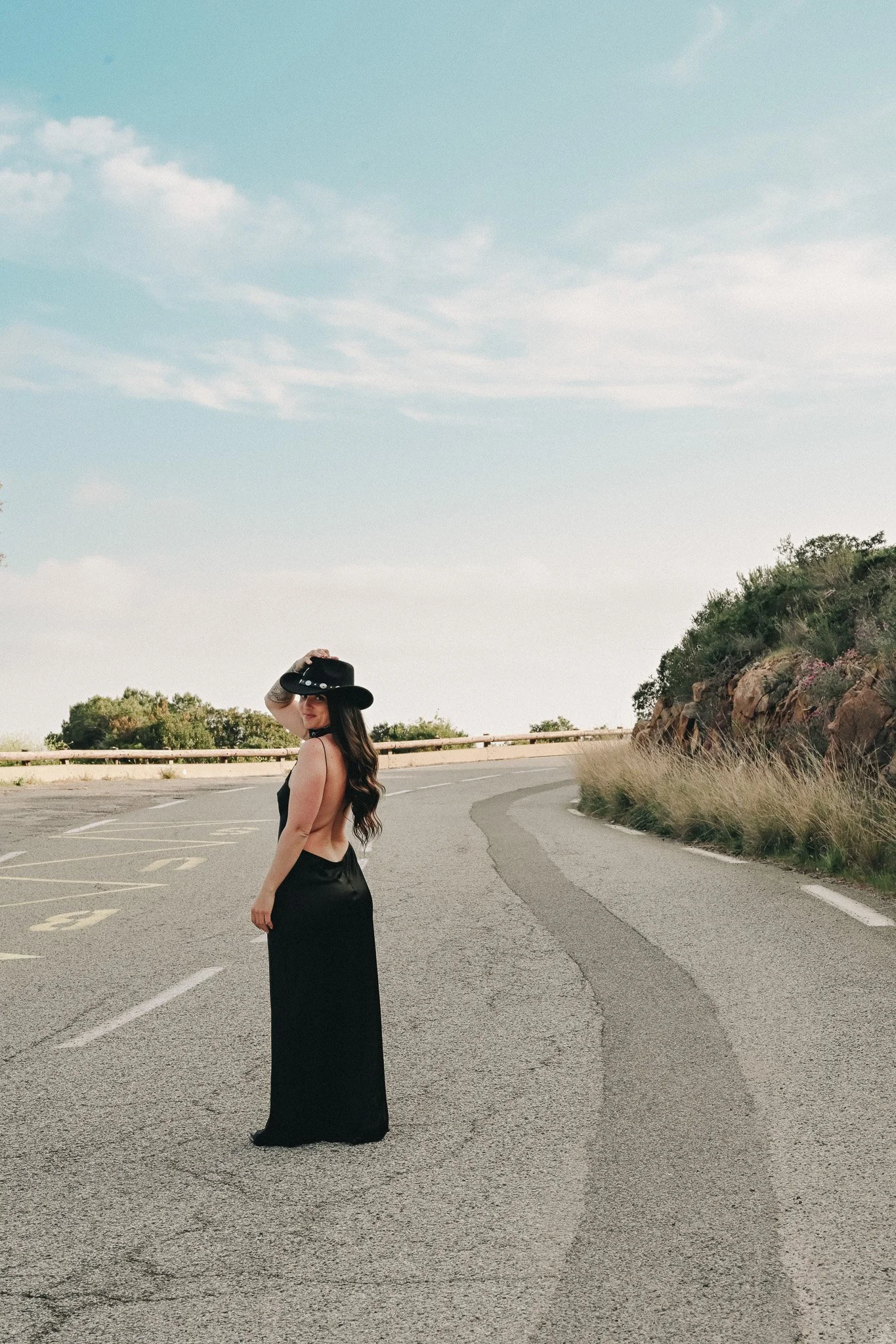 Une femme portant une robe longue noire se tient sur la chaussée d'une route, portant un chapeau noir. La scène est en plein air, avec un ciel bleu clair et des collines avec de la végétation en arrière-plan.