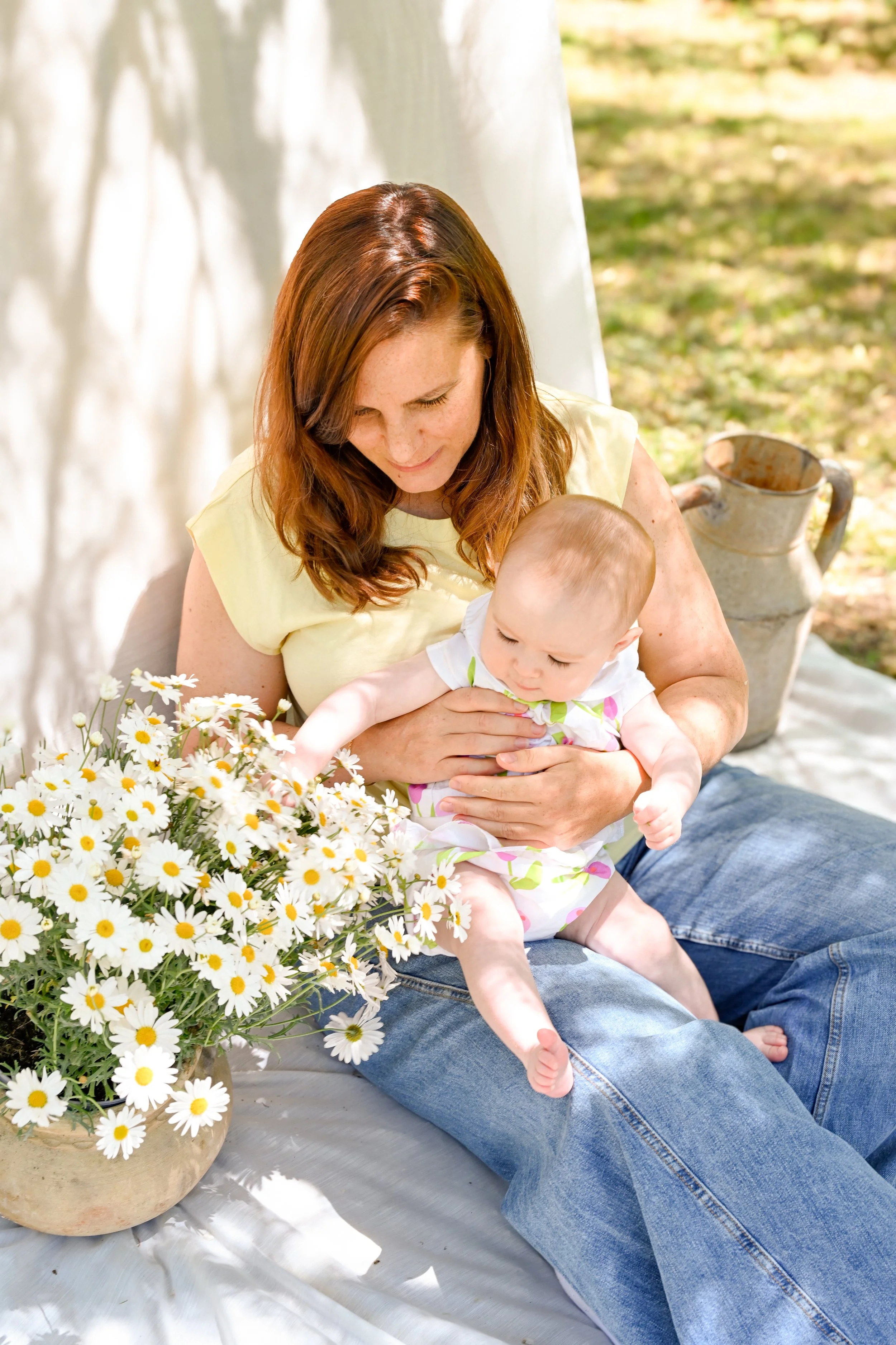 Une femme aux cheveux bruns assise sur une couverture avec un enfant, entourée de fleurs de marguerites, dans un environnement en plein air, avec un vieux pichoir en arrière-plan.