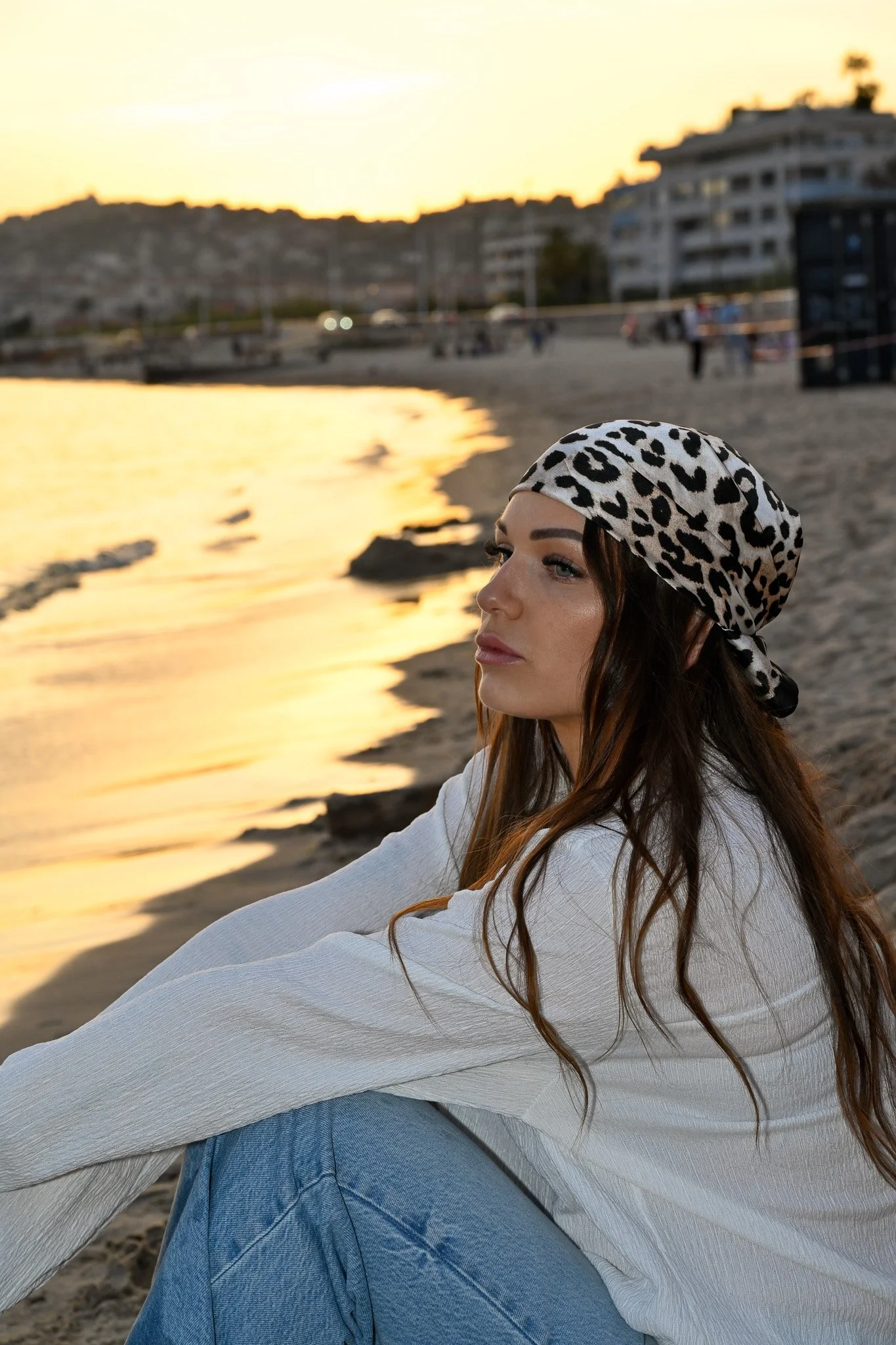 Une femme portant un bandeau léopard, vêtue d'un sweat blanc et de jeans, assise sur la plage au coucher de soleil, regardant vers la mer.