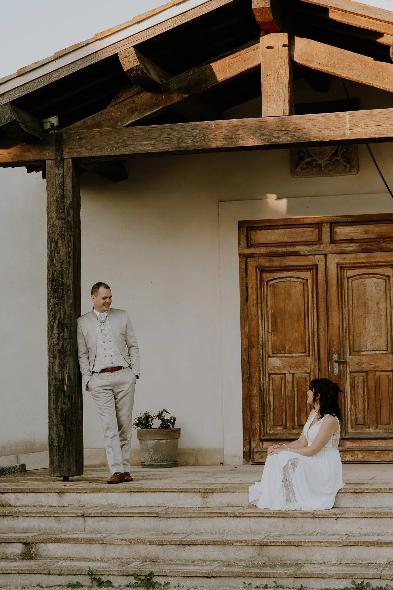 Un homme en costume blanc souriant et une femme en robe blanche assise sur des marches en pierre, devant une porte en bois, sous une structure en bois, en extérieur.